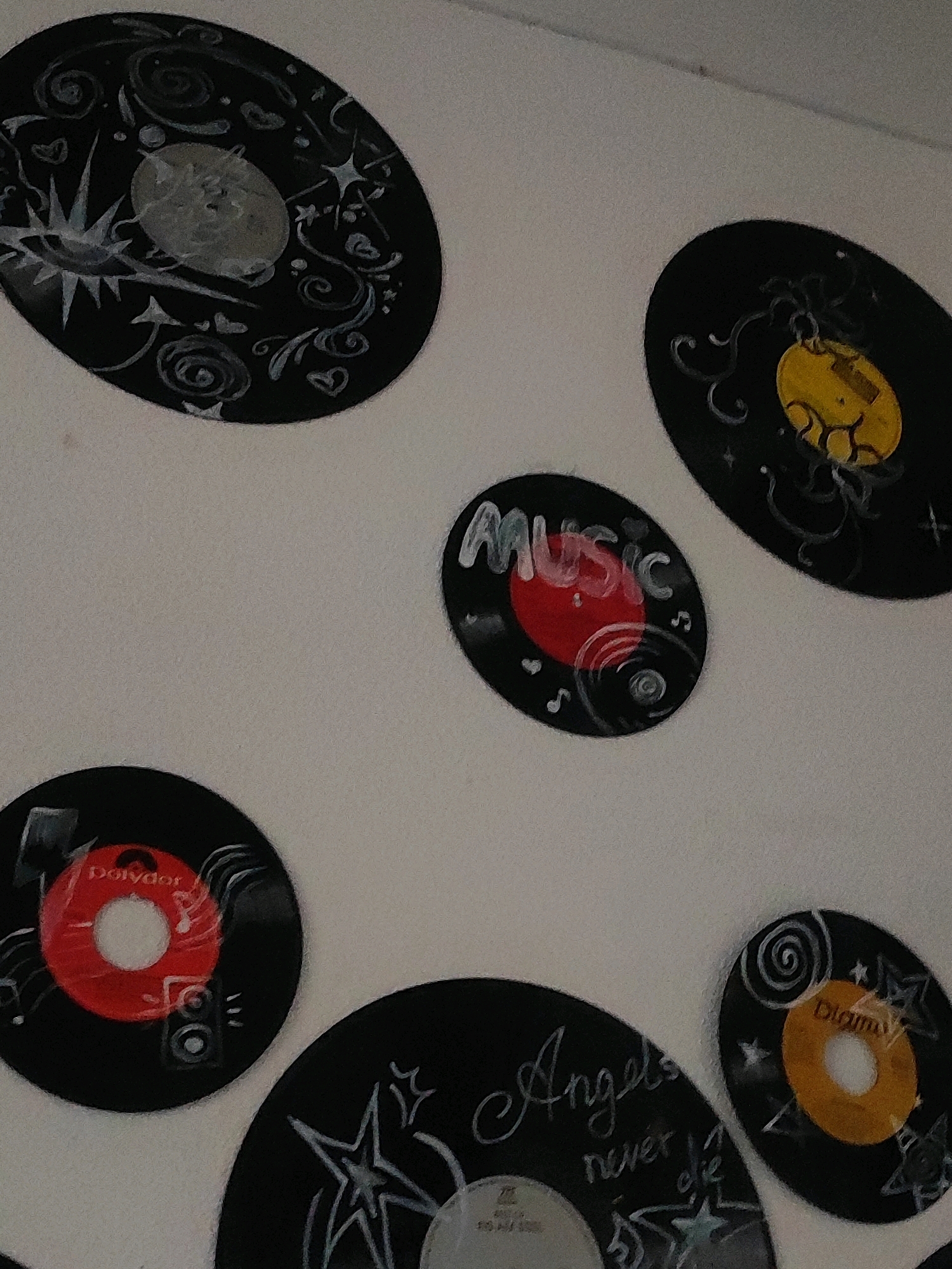 The image displays a collection of decorated vinyl records mounted on a light-colored wall. The records are of various sizes and are adorned with white and colored paint. Some records feature abstract designs, swirls, stars, and musical notes. One record is labeled "MUSIC" in white lettering with a red center. Another record has a red center with the "Polydor" logo. A small record with an orange center is also visible. The wall appears to be a plain, indoor surface, possibly in a residence or a studio. The lighting is soft, suggesting an indoor environment. The records are arranged in a somewhat scattered pattern, creating a decorative wall display. There are no people or animals visible in the image. The image does not contain any information that would indicate a specific location in Burgoberbach, Germany.