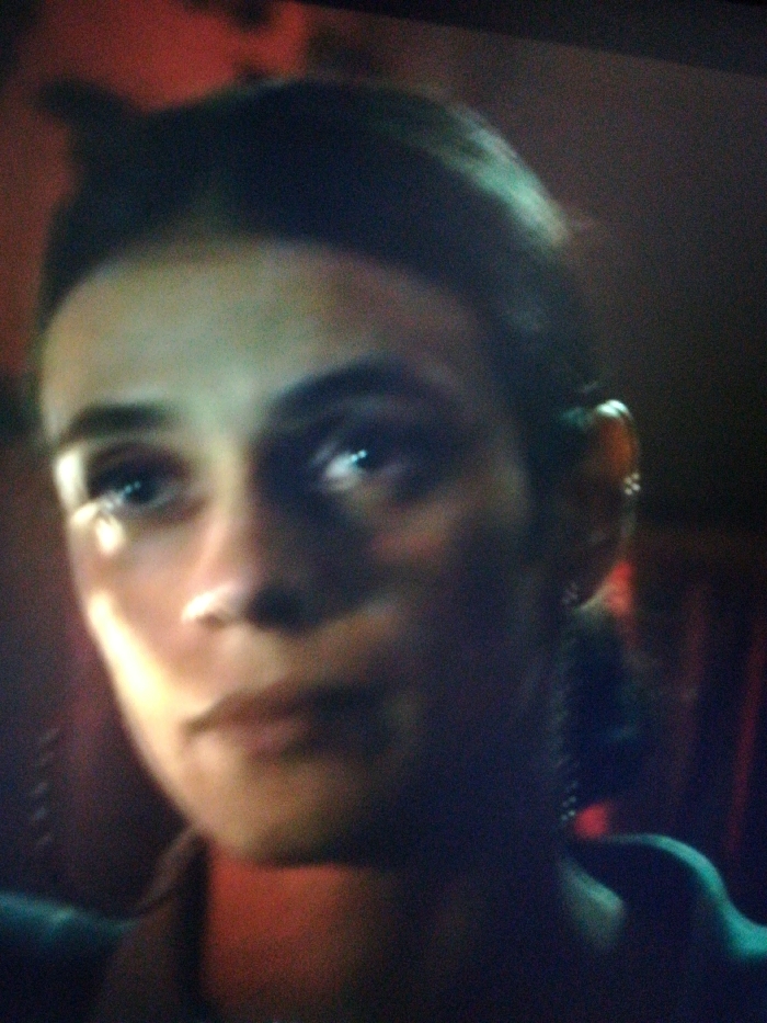 A dimly lit close-up depicts a woman's face. Her dark hair is pulled back, revealing a small earring on her left ear. Bright light reflects off her left eye. Her expression is neutral. The image has a grainy quality. A red hue is visible in the background.
