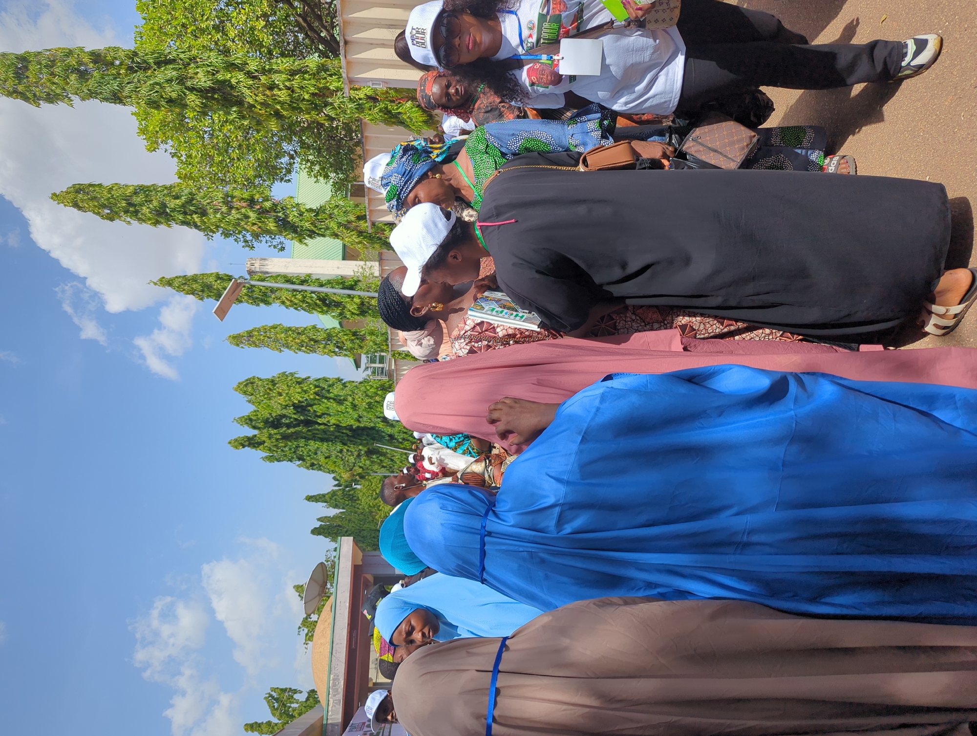 In Ungwan Liman, Nigeria, a group of people are gathered outdoors, likely at an event or gathering. The group appears to be predominantly women, some wearing head coverings like hijabs and others wearing white caps. Garments include various shades of blue, pink, black and brown. Some people are wearing white t-shirts and black trousers. The background includes trees, buildings, and a blue sky with clouds.