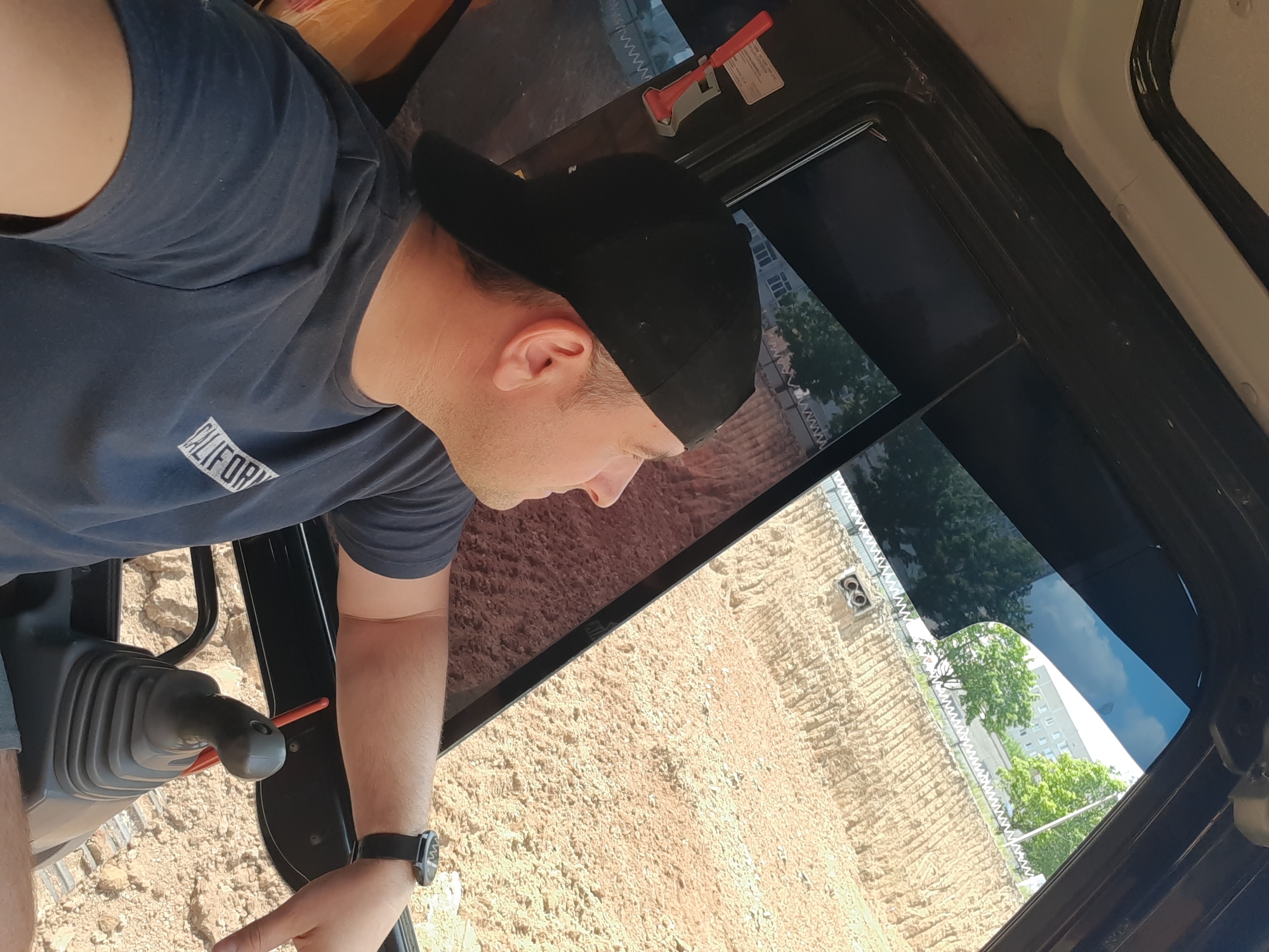A man wearing a black baseball cap and a dark blue t-shirt sits in the cabin of an excavator, looking out the window at a construction site. The excavator's control lever is visible in the foreground. Outside the window, a large pile of dirt is being worked on, with a building and trees visible in the background. The scene appears to be in Pivdennoukrainsk, Ukraine.