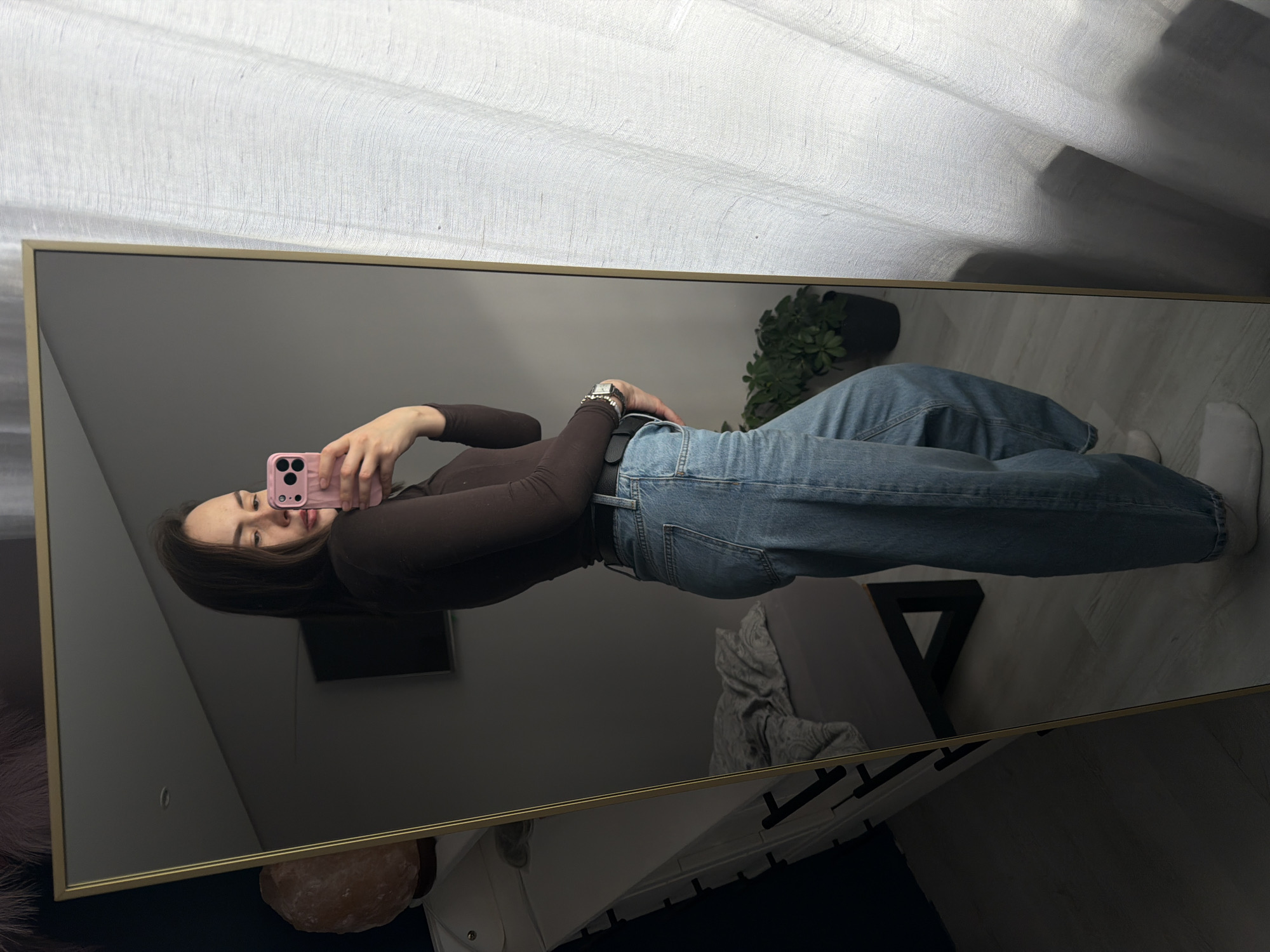 A young woman is depicted in a full-body mirror selfie, holding a pink smartphone up to her face. She has dark brown hair and a neutral expression, looking directly into the phone's camera. She is wearing a fitted dark brown long-sleeved top, loose-fitting blue jeans with a black belt, and white socks. A silver watch is visible on her left wrist.
The image is a reflection in a large, upright rectangular mirror framed in gold or brass. The background reflected in the mirror includes a light-colored floor, a potted green plant, and a dark, possibly wall-mounted, screen. Other objects reflected are a pinkish-orange salt lamp in the lower left, and what appears to be a white bed or furniture with a dark geometric pattern on its side, along with a black triangular support structure underneath.
Above the mirror's reflection, white sheer curtains are visible, suggesting a window and natural light. The setting is an indoor space, likely a home environment such as a bedroom or living room, characterized by a modern and simple aesthetic. The activity is a self-portrait being taken. No visible text is present in the image. The general time of day appears to be daytime due to the natural light. The location context provided is Chaikovsky, Russia.