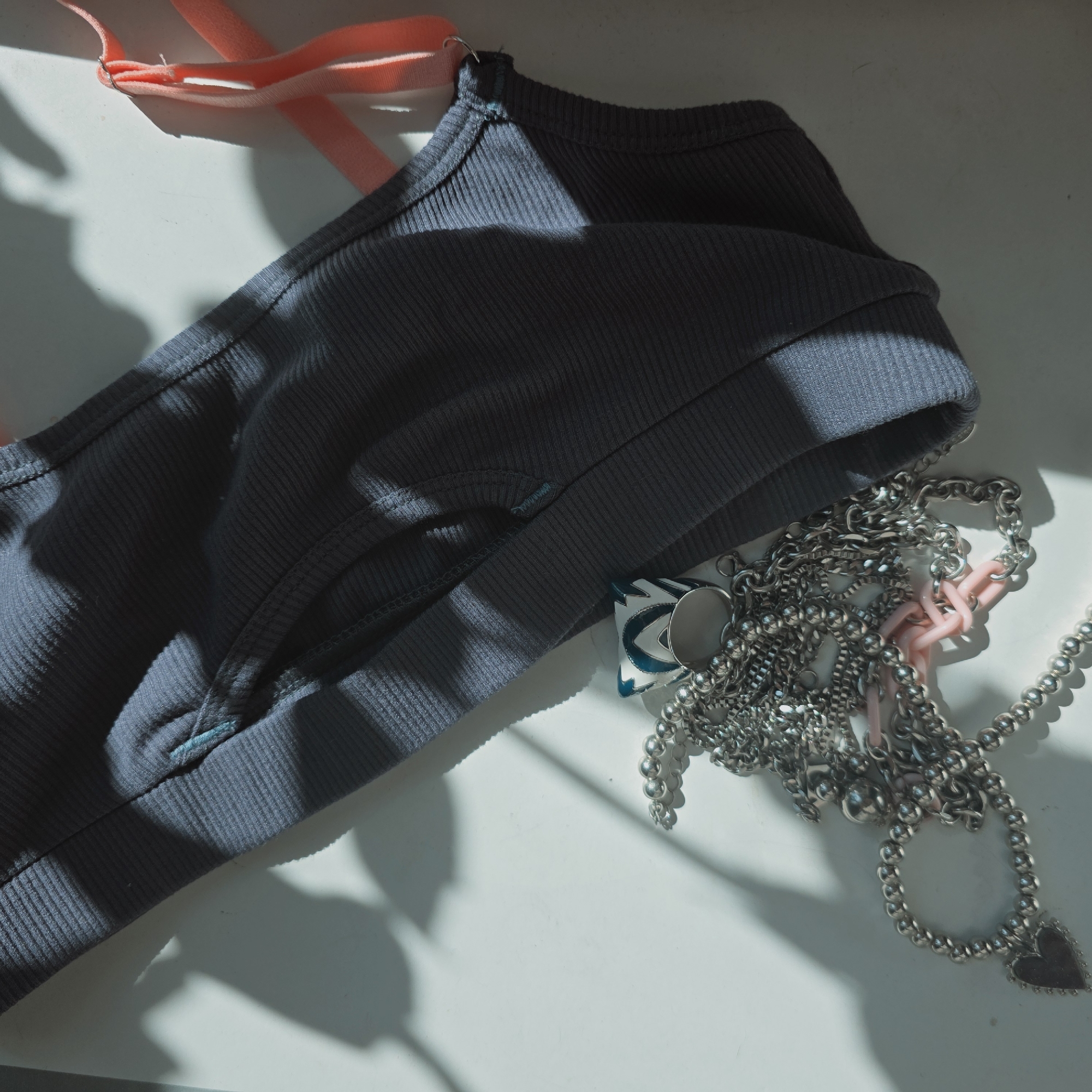 The image shows a gray ribbed top with pink straps, silver necklaces, and a blue and white accessory lying on a white surface. Shadows are cast across the scene, suggesting natural light. There is no indication of location or the city of Saint Petersburg, Russia, in the image.