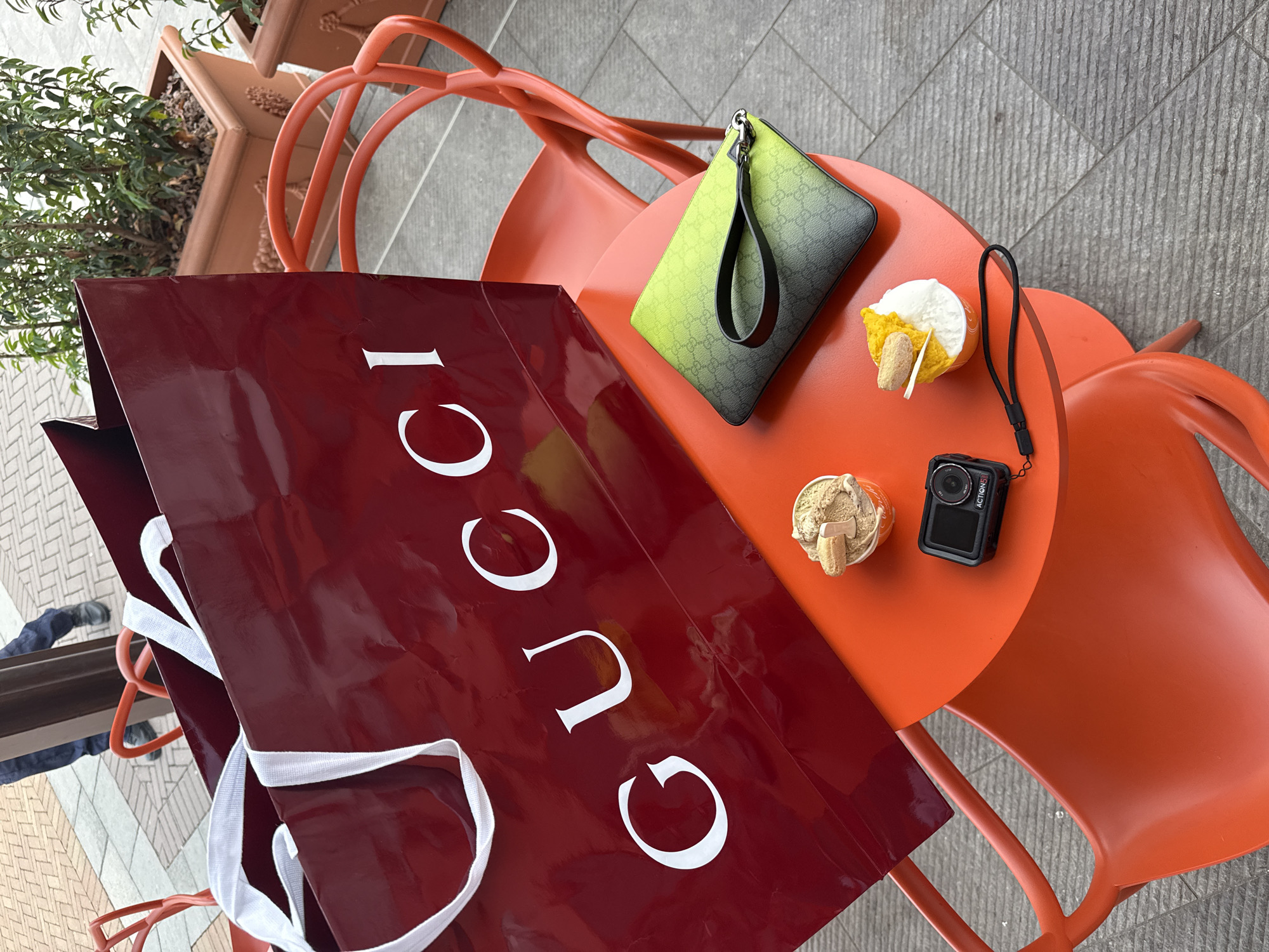 A high-angle shot shows an orange outdoor table with an adjacent plant container and gray-tiled ground. The table holds a red Gucci shopping bag, a green and black Gucci pouch, two small cups of dessert or ice cream, and a small black action camera (Action 5). The camera is in the lower-right quadrant. The items are arranged as if recently placed on the table. A person's legs are partially visible in the lower-left corner. This scene is located in Noventa di Piave, Italy.