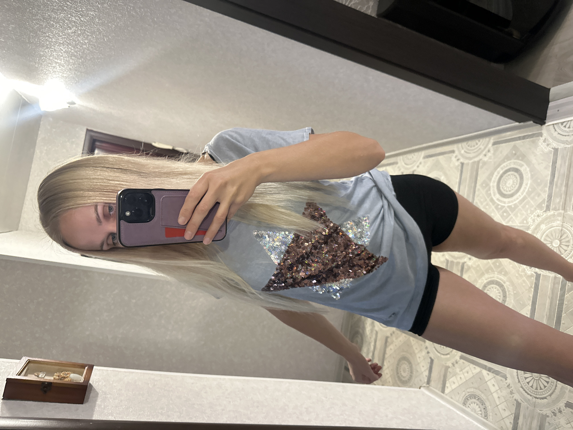 A woman with blonde hair is standing in front of a mirror, taking a selfie with her phone. She is wearing a light blue t-shirt with a sequined star design and black shorts. The room appears to be a hallway or dressing area, with light-colored walls and a patterned floor. A framed decorative item is visible on the wall to the left. The mirror reflects the woman and part of the room. The overall scene is casual and domestic. There is no specific indication of the city of Energetik, Russia within the image itself, only the provided context.