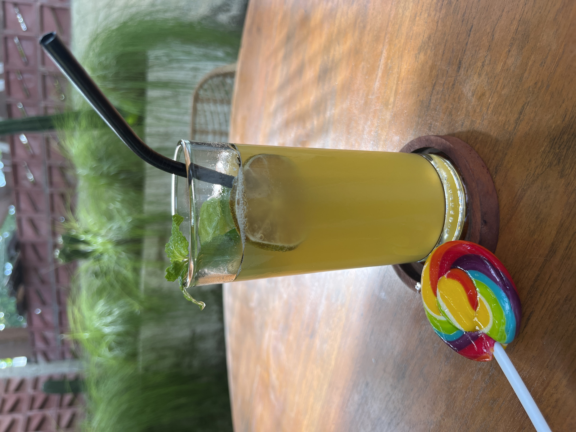 A glass of yellow liquid, likely a beverage, is placed on a wooden table. The glass contains a lime slice and mint leaves, and a black straw is inserted into it. To the left of the glass, a colorful spiral lollipop rests on the table. In the background, lush green foliage and a textured wall are visible, suggesting an outdoor or semi-outdoor setting, possibly a cafe or restaurant in a tropical location like Kuta Selatan, Indonesia. The lighting indicates it is daytime.