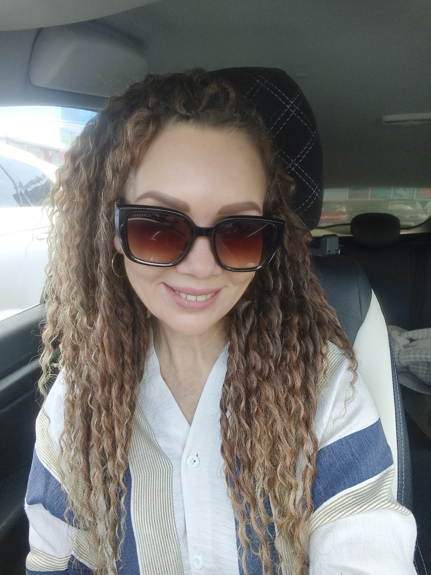 A woman is seated in the driver's seat of a car. She is wearing sunglasses and a light-colored shirt with blue and white stripes. Her curly hair is shoulder-length. The car's interior is visible, including the seats and headrests. The location is an unknown city.