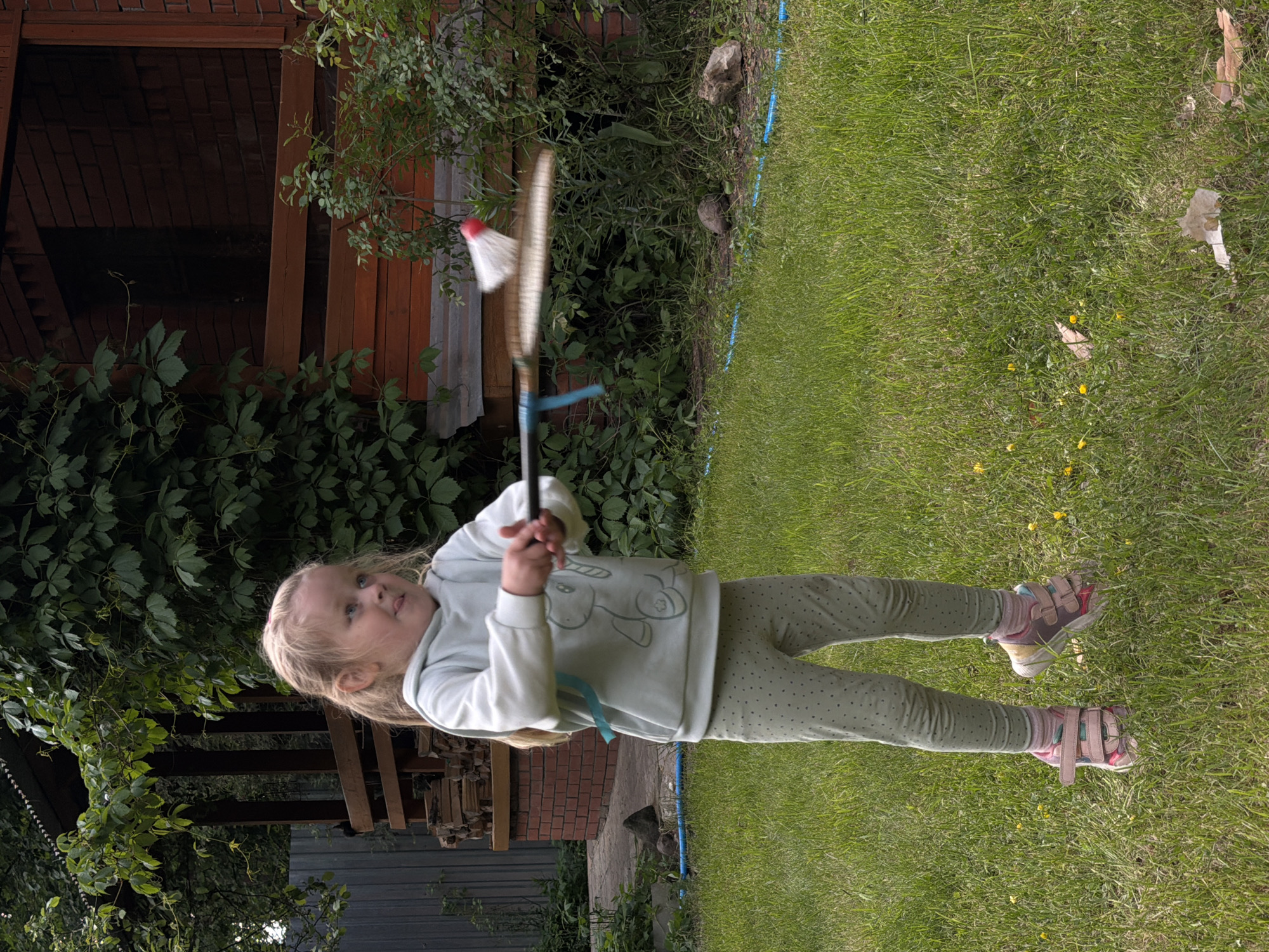 A fair-skinned young girl stands on grass, holding a badminton racket and shuttlecock. She wears a light-colored sweatshirt with a unicorn graphic, dotted green leggings, and pink sandals. Her blonde hair is pulled back. Behind her is a wooden structure, brickwork, and dense green foliage. The location appears to be a yard in Udelnaya, Russia.