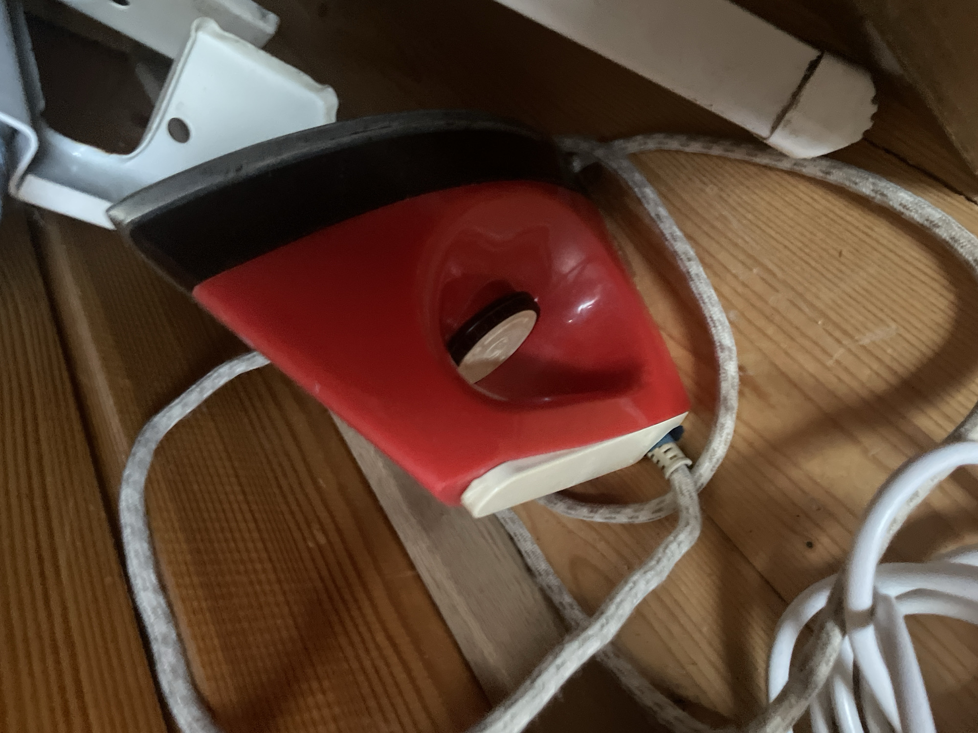 The image shows a close-up of a red and black electric iron with a white cord lying on a wooden surface. The iron is partially obscuring a white metal object. The wooden surface appears to be part of a shelf or floor. The background is dark and out of focus. There are no people or animals in the image. The overall scene suggests a storage area. The image does not provide any information that would allow for its location to be specified as being in the city of Турово, Russia.