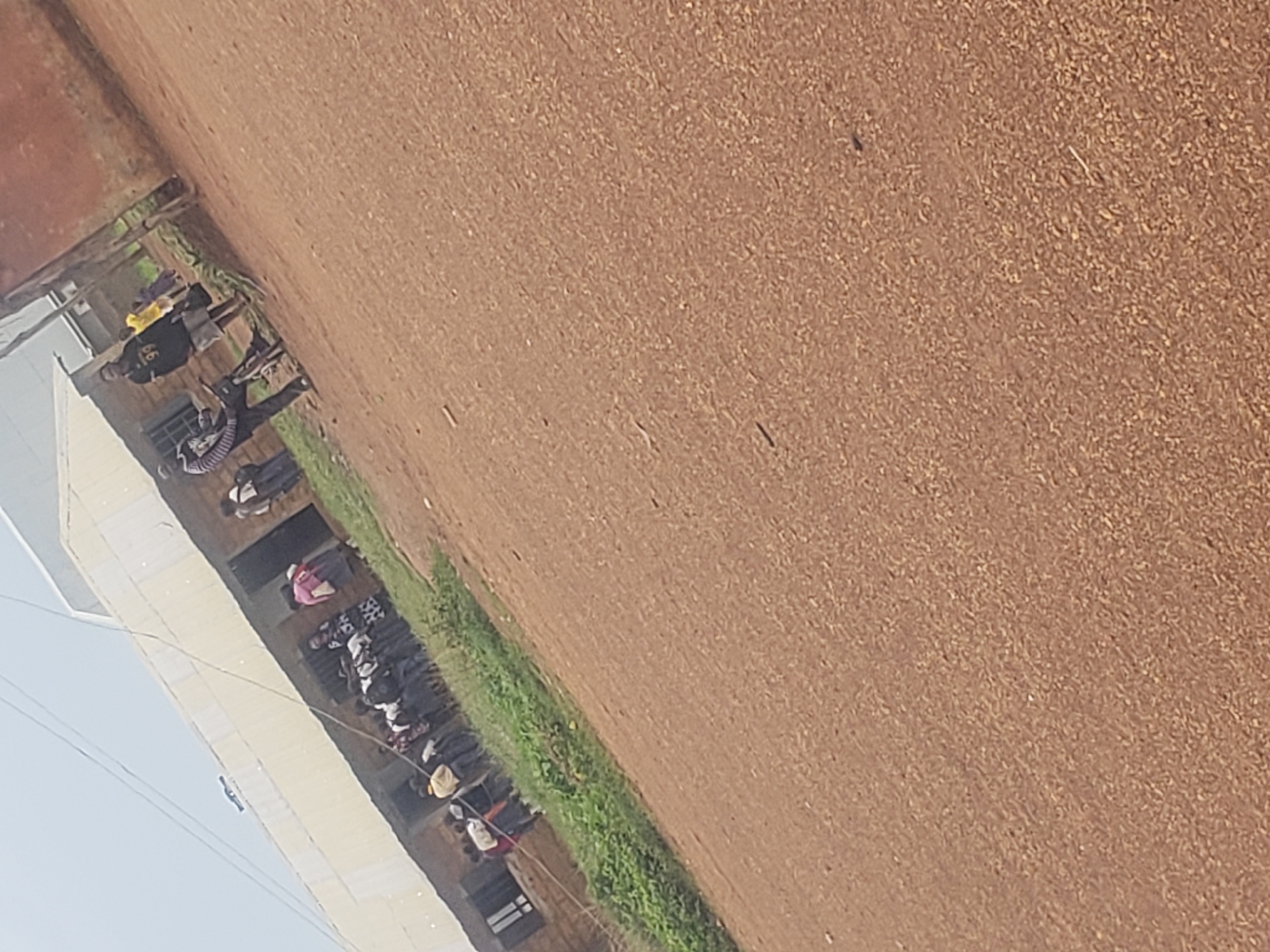 This image depicts an outdoor scene in front of a building, likely a school or community center, in Gumel, Nigeria. A group of young people, some appearing to be students, are gathered outside. One individual, dressed in a black t-shirt with the number "99" and grey shorts, stands near a bicycle. Another person in a yellow t-shirt is also visible. The ground is a reddish-brown dirt path. In the background, the building has a light-colored, possibly corrugated metal roof and brick walls. The sky is overcast. The overall impression is of a casual gathering or a moment in the daily life of the community.