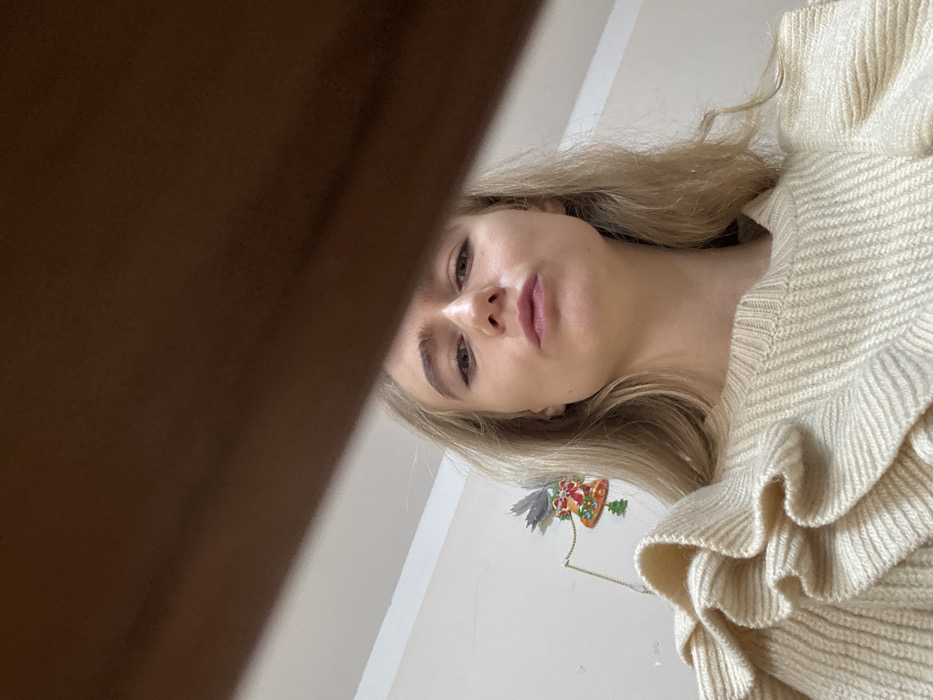 A low-angle, close-up indoor photograph depicts a female subject positioned centrally. The subject has long, blonde, wavy hair and fair skin, with her lips slightly pursed as she looks directly at the camera. She is wearing a cream-colored, ribbed knit sweater featuring prominent ruffled embellishments on the shoulders. In the background to the left of the subject's head, an off-white wall displays a Christmas-themed decoration consisting of a bell shape adorned with red ribbons and green accents, with a small green Christmas tree ornament suspended below it. The upper approximately 25% of the image is obstructed by a dark brown, linear object, consistent with a wooden beam or furniture edge, diagonally spanning from the upper left towards the lower right. The scene is located within a room in the city of Unknown, Unknown.