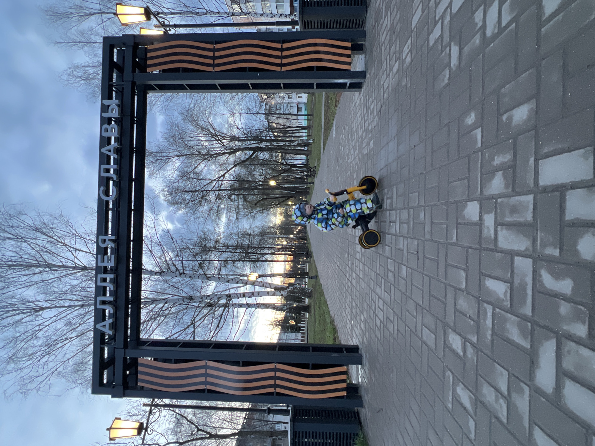 This image depicts an outdoor scene in Chaikovsky, Russia, likely a park or a memorial alley. In the foreground, a young child, dressed in a blue and green patterned jacket and a matching hat, is positioned to the right of a large archway. The child is on a small yellow scooter or tricycle, facing generally away from the camera.
The central feature is a dark metal archway that spans the paved pathway. On its horizontal crossbeam, there's a distinctive wavy pattern of black and orange stripes, resembling the St. George ribbon, a symbol of military glory and remembrance. The vertical supports of the archway bear Cyrillic text in white letters. The left side of the arch reads "АЛЛЕЯ" (Alley), and the right side reads "СЛАВЫ" (Glory), combining to form "АЛЛЕЯ СЛАВЫ" (Alley of Glory).
Beyond the archway, a long paved path stretches into the distance, lined on both sides by numerous bare deciduous trees, suggesting late autumn, winter, or early spring. Several streetlights are visible along the path, casting a warm glow, indicative of late afternoon or early evening. The sky is overcast with soft blue and grey tones. In the far background, residential buildings can be seen. The ground is covered with grey rectangular paving stones, some appearing slightly damp or reflective. The overall scene conveys a calm and serene atmosphere in a public recreational space.