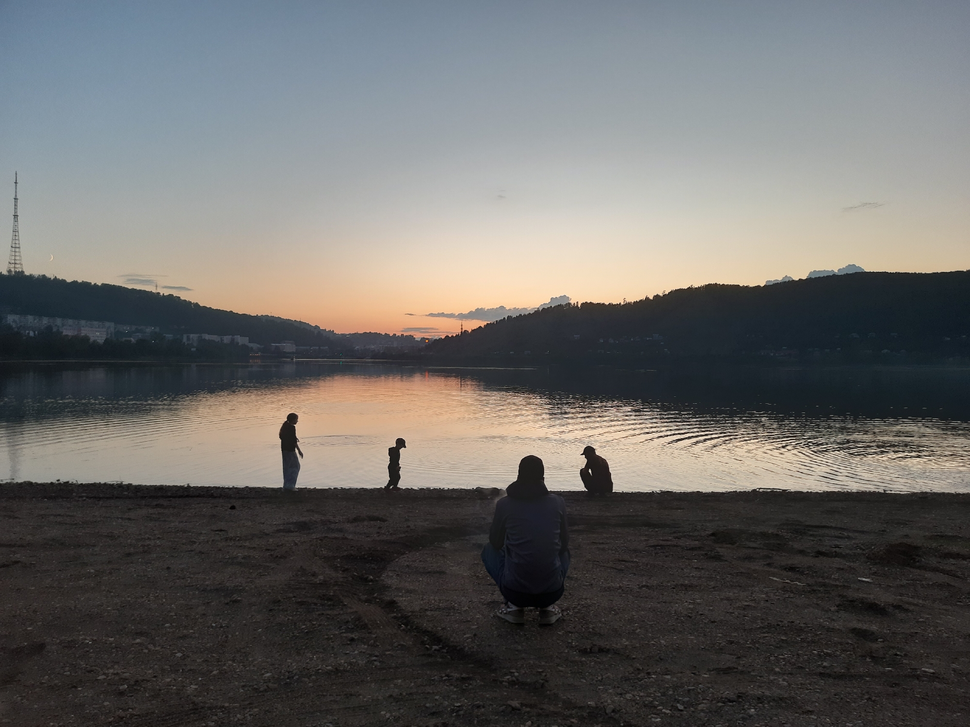 The image depicts a serene sunset over a body of water, likely a river or lake, in Zlatoust, Russia. Silhouetted against the warm hues of the setting sun, several individuals are present on the sandy shore in the foreground. One person is crouched down, seemingly observing the scene, while others stand and walk near the water's edge. The landscape is characterized by rolling hills covered in trees forming the horizon, with a city visible in the distance on the left, punctuated by a prominent radio tower. The sky transitions from a pale blue at the top to warm oranges and yellows near the horizon, indicating either dawn or dusk. The water's surface is gently rippled, reflecting the soft colors of the sky. The overall mood is tranquil and contemplative, with the silhouettes emphasizing the quiet beauty of the natural landscape and the human presence within it. There is no visible text in the image.