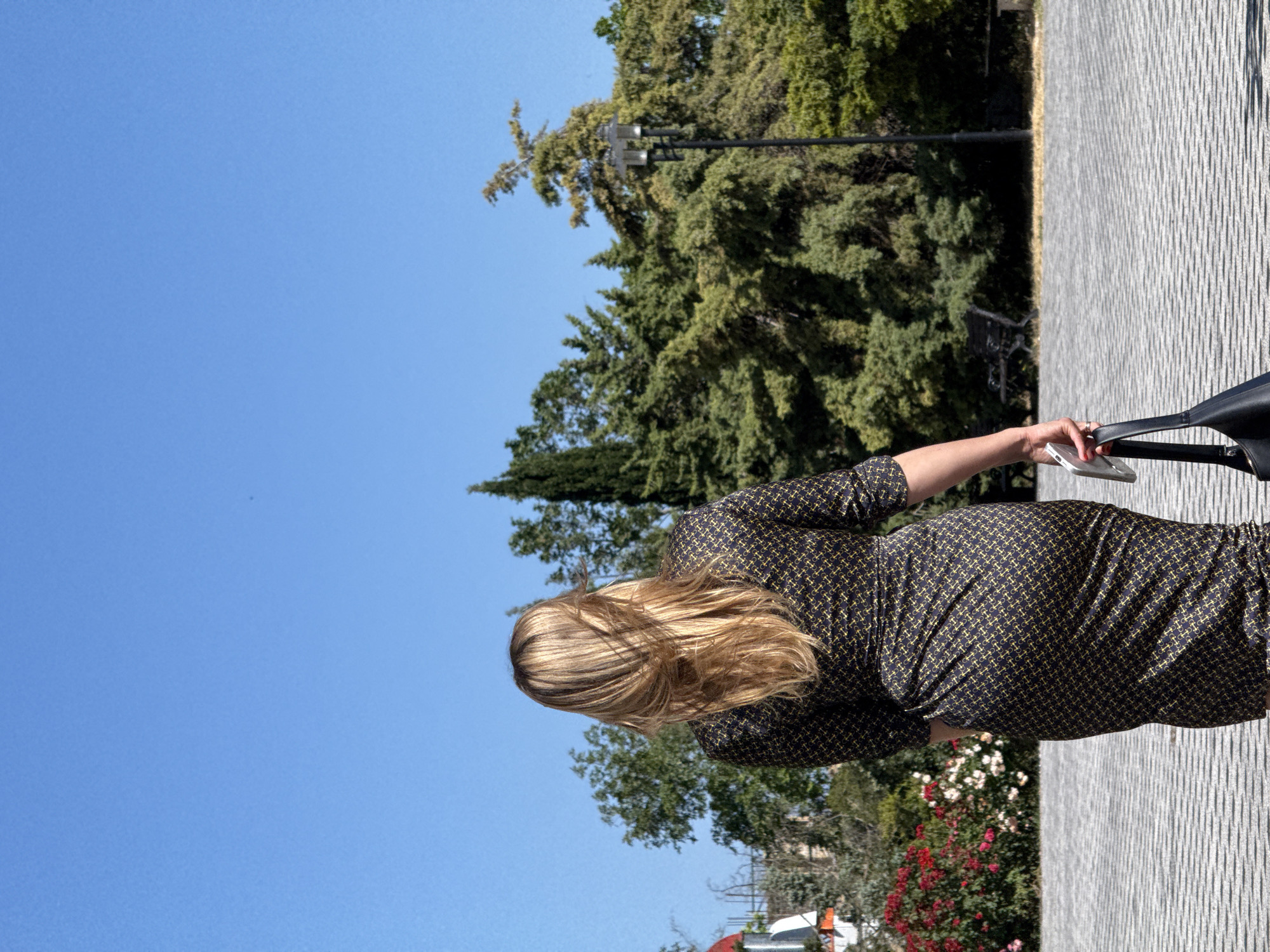 A blonde-haired woman seen from behind walks away from the camera on a paved walkway under a clear blue sky. She is wearing a dark dress with a yellow geometric pattern and carrying a black handbag in her left hand, with a silver smartphone in her right hand. Behind her, lush green trees and bushes line the background, with a lamppost visible to the right. In the distance to the left, a red and white structure is partially visible, along with a vibrant bush of red and pink flowers. The overall scene suggests a bright, sunny day in an outdoor urban or park setting.