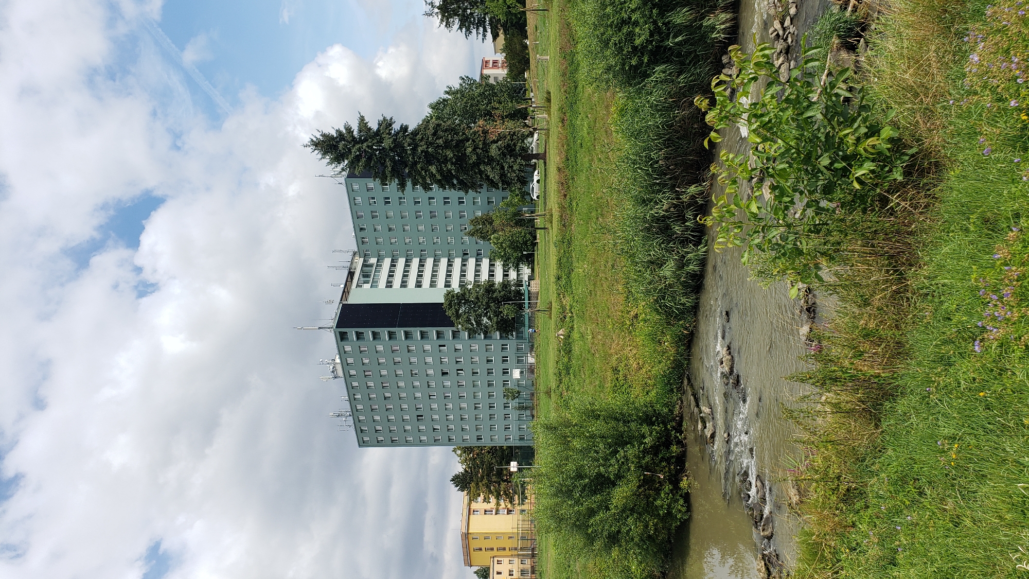 The image depicts a residential building in Prešov, Slovakia, specifically in the Sekčov housing estate. The building is a multi-story, light green structure with numerous windows and several antennas on its roof. To the left of the main building, a smaller, yellow residential building is visible. In the foreground, a river flows with rocks visible in its bed. Lush green grass and trees surround the river and the buildings. The sky is partly cloudy with some blue patches. A white car is parked in front of the main building, and a single person can be seen walking in the distance. The scene appears to be a typical urban landscape in Prešov.