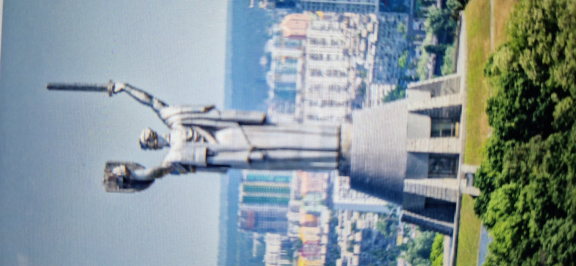 The image shows the Motherland Monument in Kyiv, Ukraine. The monument is a colossal statue of a woman holding a sword and shield, symbolizing the victory of the Soviet Union in World War II. The statue stands atop a podium, which is part of a larger complex that includes the Museum of the Great Patriotic War. The monument is located on the right bank of the Dnipro River, overlooking the city of Kyiv. In the background, the city skyline is visible, with numerous buildings and apartment complexes. The foreground is dominated by lush green trees and grass, suggesting a park-like setting. The sky is a pale blue, with no clouds visible. The image appears to be taken from a slightly elevated viewpoint, offering a panoramic view of the monument and its surroundings.