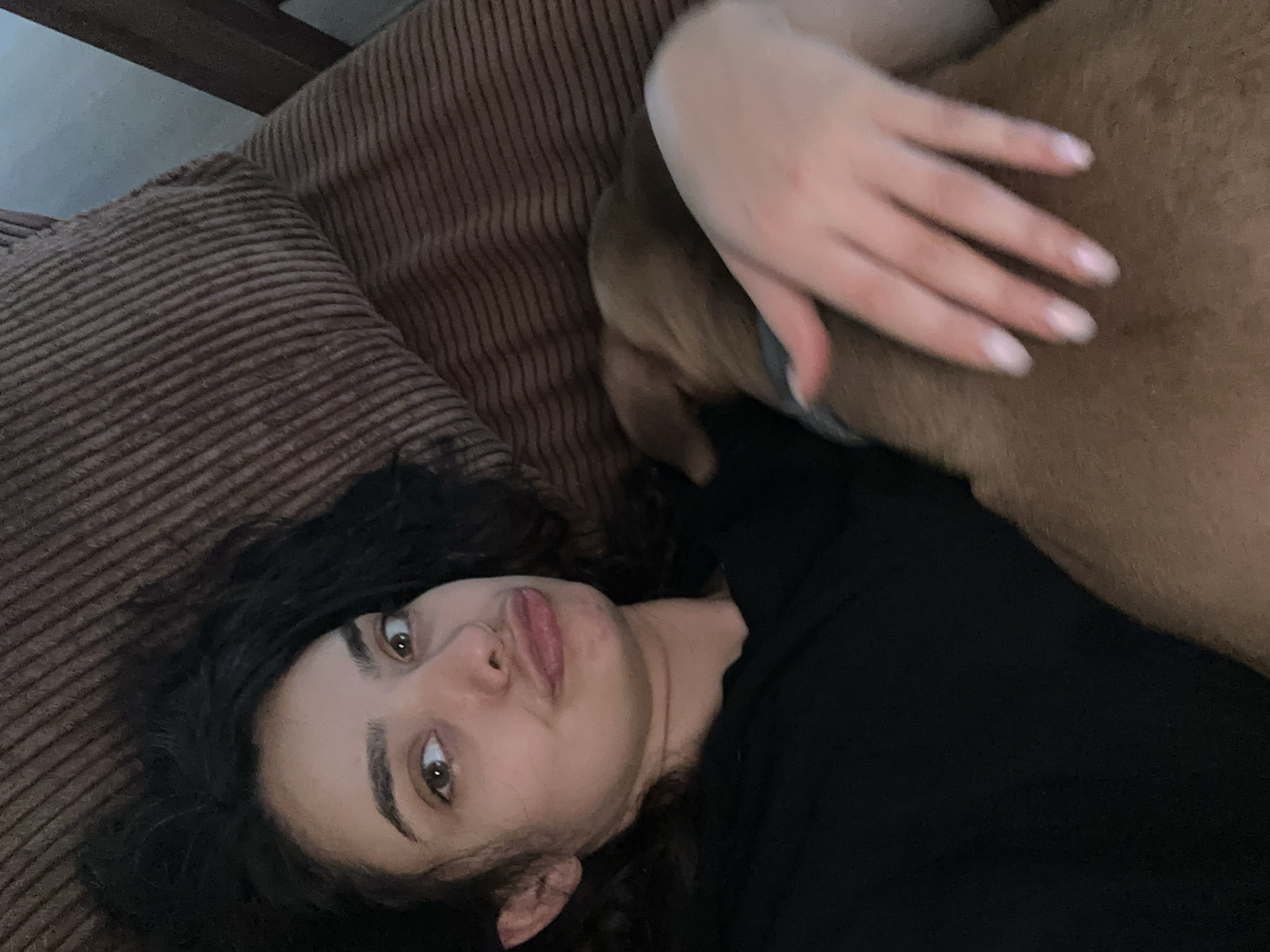 A young woman with dark, curly hair lies on a brown, corduroy sofa, looking at the camera. Her head is tilted to her left, and her mouth is pursed. Her eyes are wide open and appear to be a light brown color. She is wearing a black, long-sleeved top. A tan-colored dog is lying next to her, with its head tucked under her arm. The woman's hand is resting on the dog's back, with her fingers gently spread. The setting appears to be indoors, likely a living room, given the sofa and the presence of a wooden piece of furniture at the top left of the frame. The lighting is soft, suggesting it might be daytime or a dimly lit room. The overall mood is relaxed and intimate, indicating a moment of quiet connection between the woman and her pet. There is no visible text in the image.