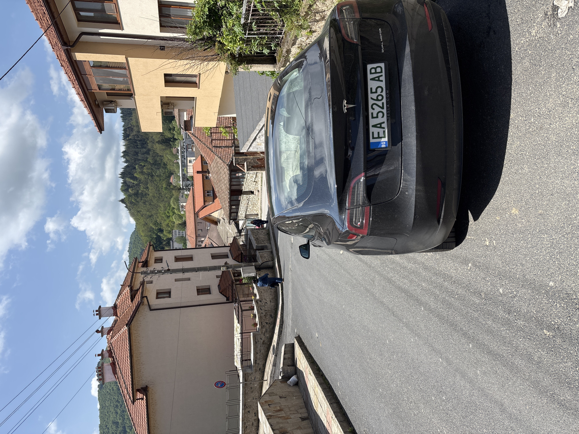 The image shows a street scene in Zlatograd, Bulgaria. A black Tesla car with license plate EA5265AB is parked on the right side of the road. The street slopes downward. Buildings with red-tiled roofs are visible on either side of the street, with some appearing to have balconies. A few people can be seen walking on the street. The sky is blue with some clouds, suggesting a sunny day. Hills covered with trees are visible in the background.