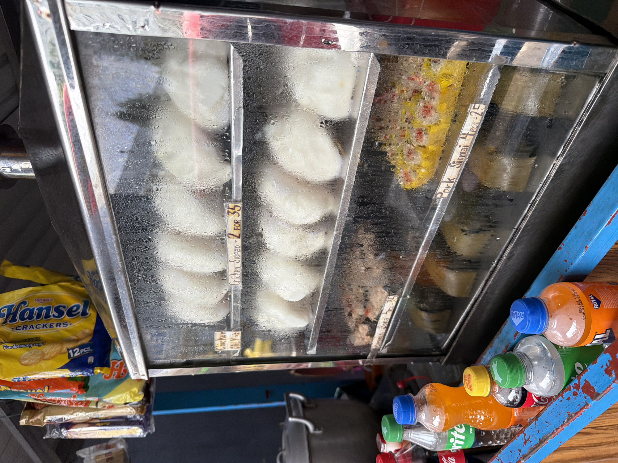 The image shows a close-up, eye-level view of a food stall, likely in Davao City, Philippines, exhibiting a variety of steamed buns and siomai displayed in a glass-fronted warming cabinet. The cabinet's glass is heavily fogged with condensation, obscuring some of the details. On the top shelf, several white steamed buns are visible. Below them, on the second shelf, are more of these white buns. The third shelf holds what appear to be small, prepared dishes with visible ingredients, possibly another type of siomai or dumplings. The bottom shelf displays a colorful arrangement of what is identifiable as pork siomai, with a handwritten label indicating "Pork Siomai 4 pcs - 25". Adjacent to the warming cabinet, on the left, are several plastic bottles of beverages, including what appears to be a bottle of Sprite and an orange-colored soda, positioned in a blue-painted wooden holder. Behind the bottles, a large metal pot with handles is partially visible. In the background, hanging bags of snack crackers, specifically "Hansel Crackers," are evident. The overall environment suggests an outdoor or semi-outdoor food vendor setting.