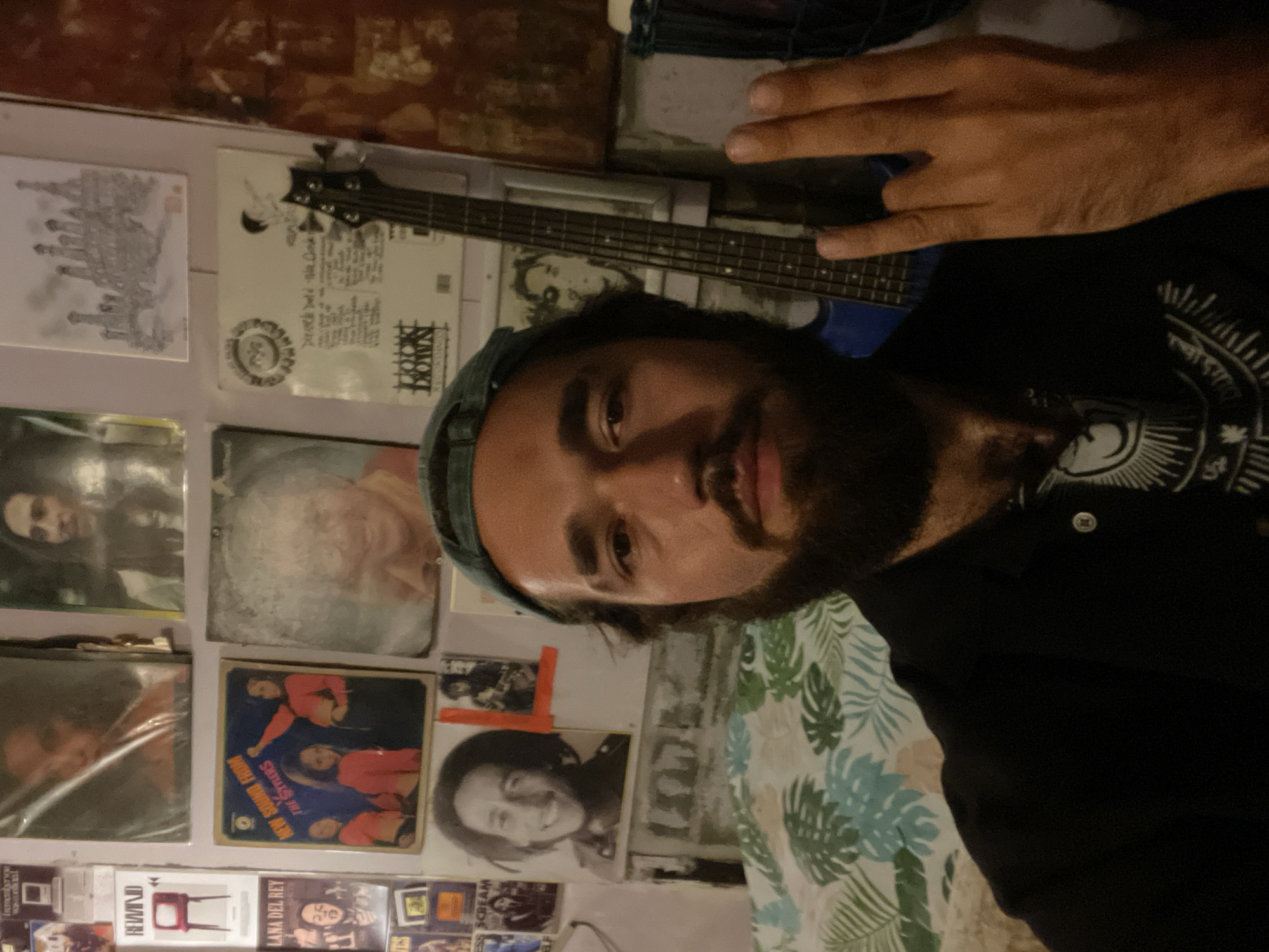The image is a close-up, vertically oriented selfie of a person with dark hair and a beard, wearing a blue baseball cap and a black shirt with a white graphic design. Their right hand is visible, with two fingers extended upwards in a peace sign. The person is looking directly at the camera with a neutral expression.
Behind the person, a wall is covered with various items. There is a bass guitar with a white pickguard and dark fretboard resting horizontally. Below the guitar are several album covers and posters. One album cover, featuring a group of people in red shirts against a blue background, has text that appears to read "THE STYLES NEW SOUND ALBUM." Another poster is a black and white image of Bob Marley. Further down, other album covers and posters are visible, including one with "RE-WIND" and a red television set, and another with "LANA DEL REY." The wall itself appears to be a light color, possibly painted or wallpapered. To the right of the person, a patterned fabric with green and blue leaves is visible. The lighting suggests an indoor setting, possibly with ambient or artificial light. The overall scene has a casual, informal, and possibly music-themed atmosphere. Given the location context of Chalong, Thailand, the setting is likely a room or a studio in that area.