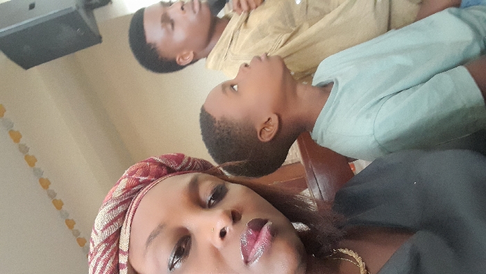 The image displays an indoor scene in Badore, Nigeria, featuring three individuals. In the immediate foreground, positioned to the left, is an adult female with dark skin, wearing a patterned headwrap with red, gold, and light-colored stripes, and a dark-colored top. A gold-toned necklace is visible, and she has a nose piercing on her left nostril. She is looking directly towards the camera. To her right, a young male child with dark skin and short dark hair is seated, wearing a light blue or mint green t-shirt. He is depicted in a profile view, looking upwards and to his right. Further in the background, to the right of the child, an adult male with dark skin and short dark hair is visible, wearing a light brown long-sleeved shirt. His eyes are closed, and he is looking downwards. All individuals appear to be seated, with a dark brown wooden bench or pew partially visible in the middle ground. The background consists of light-colored walls adorned with a garland of yellow and light-colored decorative cutouts strung along the ceiling line. A black rectangular speaker is mounted on the wall in the upper right portion of the frame.