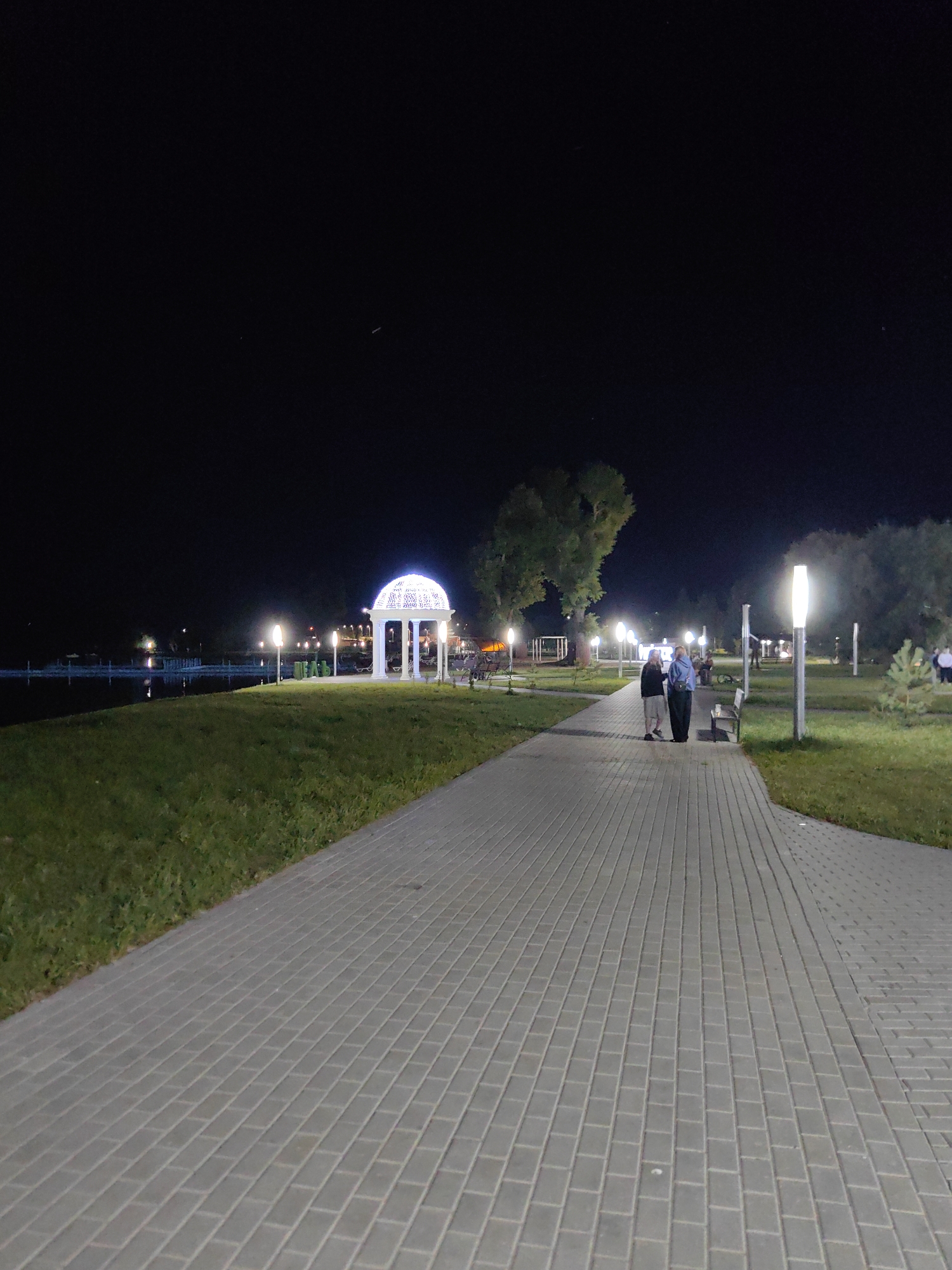 This is a nighttime photograph of a paved walkway in a park or recreational area. A white, ornate gazebo with illuminated decorative lights is visible on the left side of the image, situated on a grassy area adjacent to a body of water. Further along the walkway, two individuals are standing and conversing. The pathway is lined with modern, vertical lampposts, and trees are present in the background. The scene appears to be located in Myadzyel, Belarus.