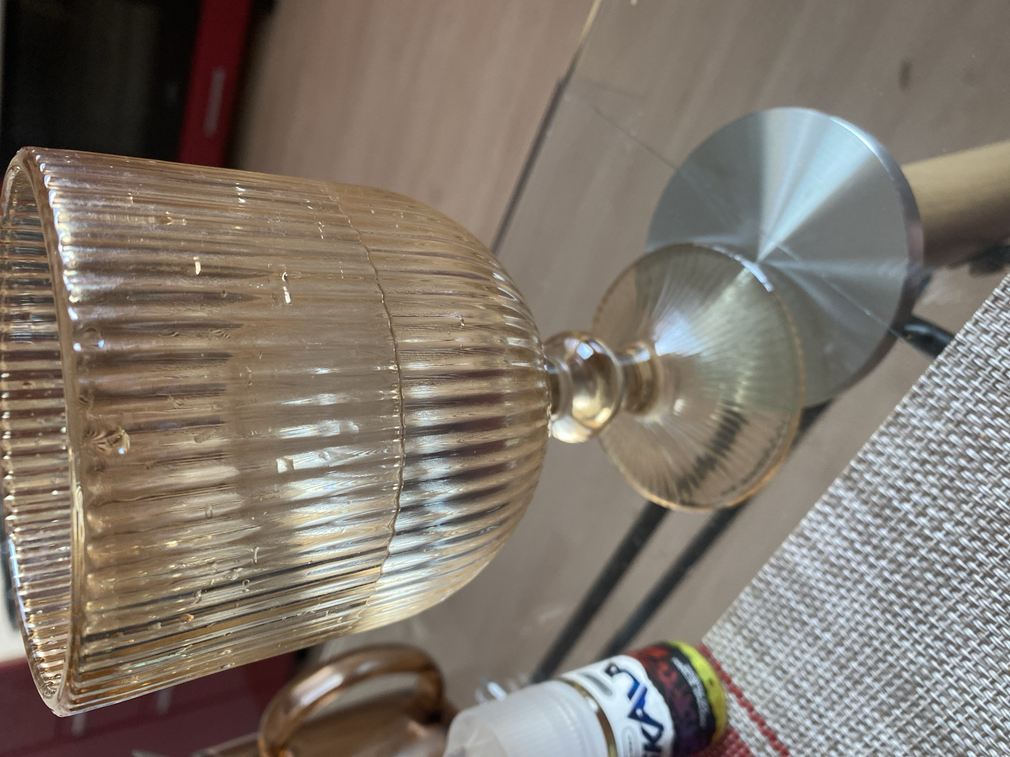 The image displays a close-up of an amber-colored, ribbed glass goblet resting on a transparent surface, likely a glass table, in an indoor setting in Yekaterinburg, Russia. The goblet's body and stem are characterized by prominent vertical fluting or ribbing, which creates intricate light refractions and reflections. Its base is a circular, highly reflective, silver-toned metal disk, further enhancing the play of light.
Through the clear tabletop, a woven placemat or textile with a brown and white crisscross pattern is visible on the right. Below the table, towards the bottom right, a small bottle with a colorful label is partially obscured. Visible text on the bottle's label includes "BLACK LABEL" in white letters on a dark background, along with a stylized logo that appears to contain letters such as "A" or "K", and other unreadable text. The lighting suggests an indoor environment. No people are present, and no specific activity is taking place; the goblet appears to be stationary, either as a decorative item or awaiting use.