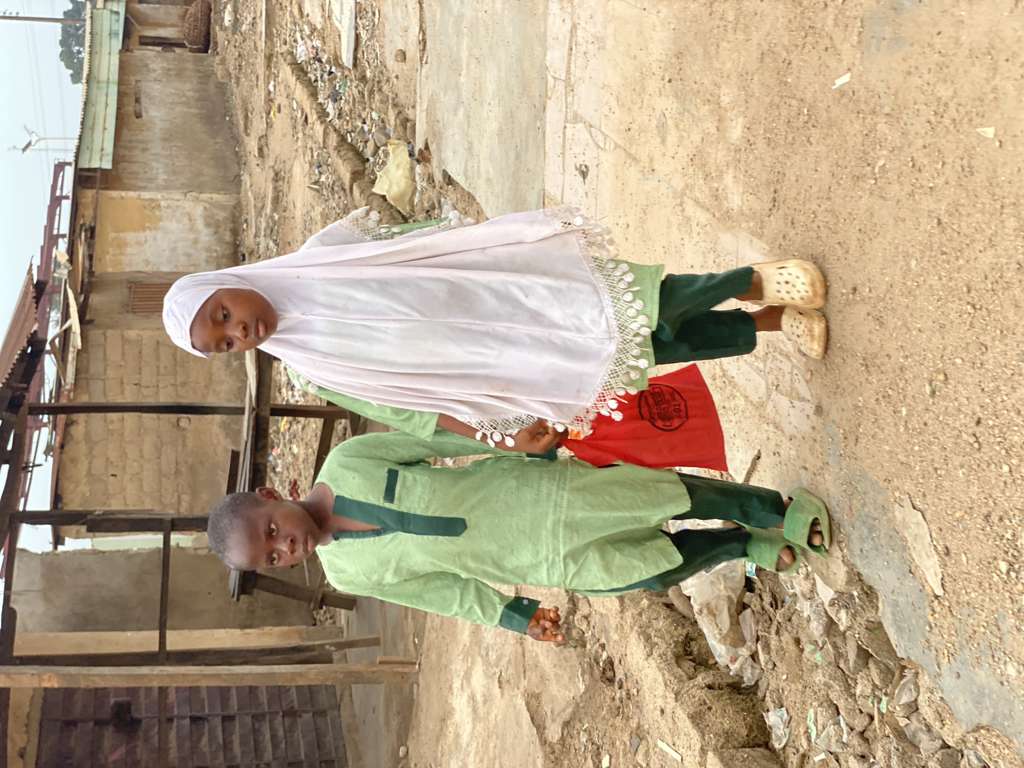 Two children, a boy and a girl, are standing in what appears to be an impoverished area in Kafanchan B, Nigeria. The girl wears a white hijab, a green dress, and light-colored clogs. The boy is wearing a green tunic-style shirt and pants with green sandals. They are standing on a rough dirt and concrete surface. Behind them is a dilapidated structure with a partially collapsed roof and exposed wooden and metal beams. Litter and debris are scattered around the scene. The overall environment suggests a low-income or slum area.