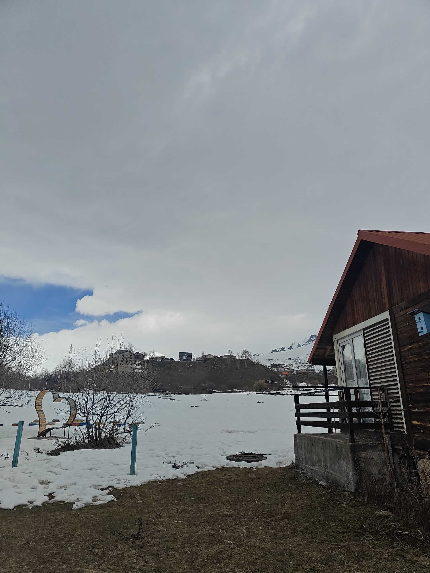 The image depicts a serene winter landscape featuring a rustic wooden cabin on the right, set against a backdrop of snow-covered ground and distant hills. The cabin has a red A-frame roof, wooden siding, and a small balcony with dark railings. A light blue birdhouse is attached to its exterior.
The ground in the mid-ground is covered in melting snow, revealing patches of brown grass and earth. On the left side of this snowy expanse, a large, ornate wooden sculpture or bench shaped like a heart is visible, surrounded by bare winter trees and shrubs. Two teal-colored posts are planted in the ground nearby.
In the mid-distance, hills or mountains rise, topped with scattered buildings, including what appears to be a partially built concrete structure and several smaller houses. These structures are nestled among bare trees. Further in the background, snow-capped peaks are faintly visible beneath the sky.
The sky is predominantly overcast with heavy grey clouds, but a striking patch of bright blue sky with white, fluffy clouds breaks through on the left side, offering a glimpse of clearer weather. The overall impression is one of tranquil solitude during a cold season. The time of day appears to be daytime, characterized by soft, diffused lighting. No human activity or individuals are visible, and there is no discernible text in the image.