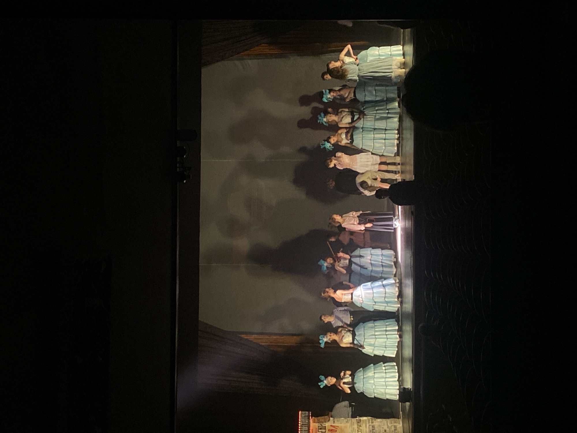The image shows a stage performance with a group of performers in period attire, including dresses and headwear, likely in a theater or auditorium. The stage setting includes backdrop and props, such as a newspaper stand, creating an atmosphere evocative of historical or literary themes. Some members of the ensemble are engaged in various actions. The scene, in Rusanovo, Russia, is illuminated by stage lighting.