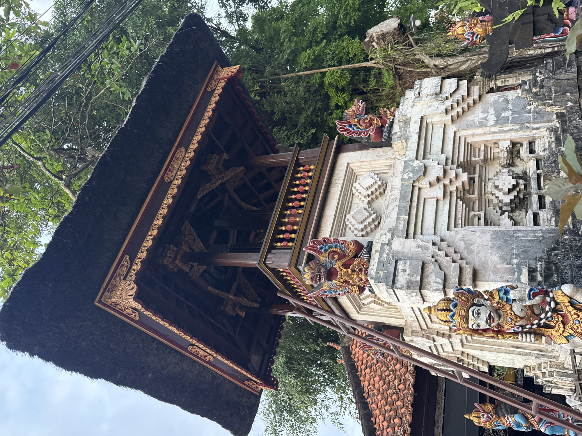 The image shows a Balinese Hindu temple structure in Denpasar, Indonesia. The structure has a traditional thatched roof and intricate carved details with colorful accents. The temple's tiered design is apparent with ornamental figures adorning its façade. The environment consists of lush greenery visible around the structure, implying a tropical setting. The image appears to be taken from a low angle, showcasing the temple's height and architectural details.