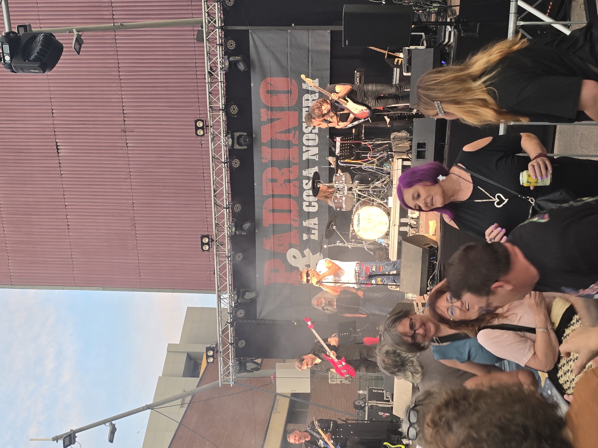 The image captures an outdoor live music event in Cornellà de Llobregat, Spain, featuring a band performing on a temporary stage while an audience observes from the foreground. The scene is set in an urban environment, likely a public square or street, against the backdrop of a large building with a corrugated reddish-brown facade.
The stage, centrally positioned, features a metal trussing structure adorned with numerous stage lights and moving head lights. A prominent black banner serves as the stage backdrop, displaying the band's name "PADRINO & LA COSA NOSTRA" in distressed red and white lettering. Five male musicians are performing on stage. From left to right: a man with grey hair, wearing a black studded outfit, plays an electric guitar; a man with dark hair and a beard, in a black shirt, plays a bright pink/red bass guitar; a man with long grey hair and a beard, in a black t-shirt, stands near a microphone; the lead vocalist, wearing a white short-sleeved shirt, sunglasses, and patched jeans, sings into a microphone; and a person with long hair plays a light brown electric guitar, with a drum kit (showing "Roland" branding on the bass drum) partially visible behind them. Various amplifiers, speakers, and cables are arranged on the stage floor, and atmospheric smoke or haze is present.
In the foreground, a diverse group of audience members is gathered. Notable individuals include: a woman with short, vibrant purple hair, wearing a black top with cut-out shoulders and a heart-shaped pendant, holding a light-colored beverage can; a woman with long blonde/light brown hair, wearing a black t-shirt and a silver hair clip, viewed from the back; a woman with dark grey hair and glasses, wearing a blue-green top, smiling; a woman with reddish-brown hair and glasses, in a light pink top, holding a patterned bag, laughing; and a man with short dark hair, wearing a black t-shirt, seen in profile, engaging with the women. Other individuals are partially visible in the extreme foreground.
Beyond the main building behind the stage, other multi-story buildings with lighter grey and beige facades are visible under a partly cloudy, light blue sky. Streetlights and utility poles are also present in the peripheral background, confirming the urban setting. The overall lighting suggests either late afternoon or early evening, with stage lights already activated to enhance the performance.