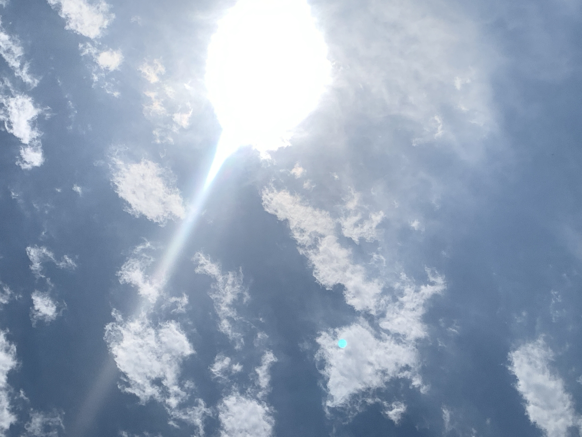 The image shows a bright, sunlit sky with scattered white clouds. The sun itself is a brilliant, overexposed orb in the upper right quadrant, casting a strong glare that extends diagonally across the frame. A faint rainbow or lens flare effect is visible emanating from the sun. In the lower left quadrant, there is a small, turquoise-colored orb, likely a photographic artifact or reflection. The sky is a clear blue, suggesting an outdoor daytime scene. The image does not contain any identifiable landmarks or structures that would indicate a specific location within Khazar Raion, Azerbaijan.