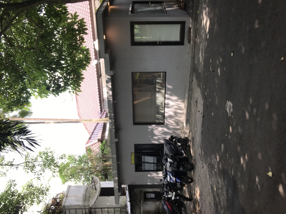 The image shows a small, grey building with a yellow sign that reads "Musholla" on the wall. There are two windows and a door on the front of the building. The windows have black frames and blinds. The door is also black and has a small window on the top. The building is located on a paved driveway with a few motorbikes parked in front. The driveway is shaded by a large tree with green leaves. The location is in Malang, Indonesia, at the coordinates -7.98 latitude and 112.617 longitude. There is a red-roofed building visible in the background behind the tree.