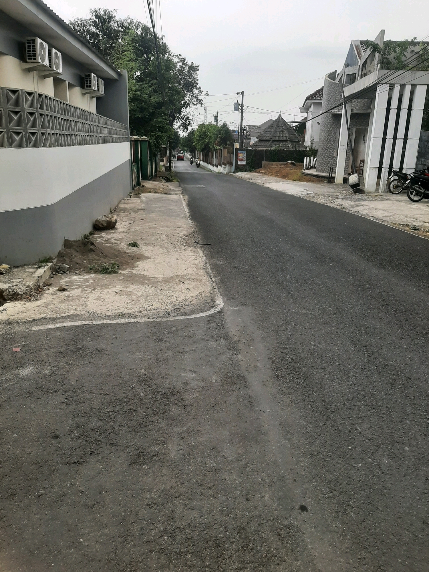 The image captures a street scene in Sumampir, Indonesia. The perspective is from ground level, looking down a paved asphalt road that curves slightly to the right. On the left side of the road, a grey and white building with multiple air conditioning units is visible, bordered by a decorative patterned wall. A narrow, worn concrete sidewalk runs alongside this building. Further down the road on the left, a green gate is visible, and beyond that, trees and distant houses. On the right side of the road, a modern-looking building with distinctive white vertical columns and a curved stone facade is present. Two motorcycles are parked on the sidewalk in front of this building. The sky is overcast. The road appears to be in moderate use, with no people immediately visible in the foreground.