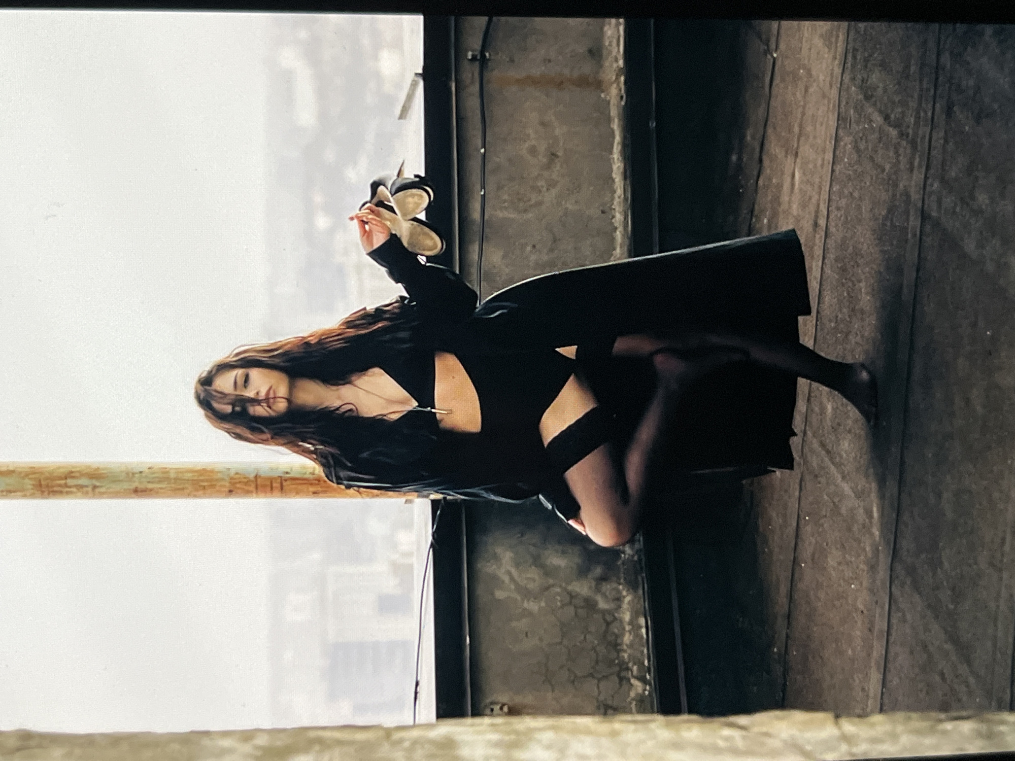 A young woman with long, dark hair stands on a rooftop in front of a hazy cityscape, holding a pair of white high-heeled shoes in her right hand. She is wearing a black lingerie set consisting of a bra and high-waisted briefs with thigh-high stockings, and an open black trench coat. Her left leg is bent at the knee, and her right leg is extended slightly forward. The woman is looking directly at the camera with a serious expression. The rooftop appears to be aged, with concrete walls and a weathered wooden floor. A rusty metal pole is visible behind her to the left. The background shows a blurred view of buildings under an overcast sky.