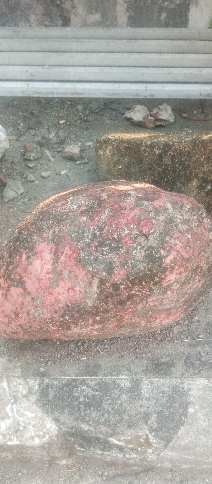 An oblong, reddish-brown object, possibly a large rock or a discarded piece of equipment, rests on a rough, greyish surface. The object is weathered and appears to have patches of green moss or lichen on its surface. It is positioned in front of a larger, darker, textured object that is partially obscured by shadows. In the background, several grey, cylindrical objects are stacked horizontally. The ground around the objects is composed of small stones and debris. This scene appears to be an outdoor location, potentially in an industrial or construction area within Kediri City, Indonesia.