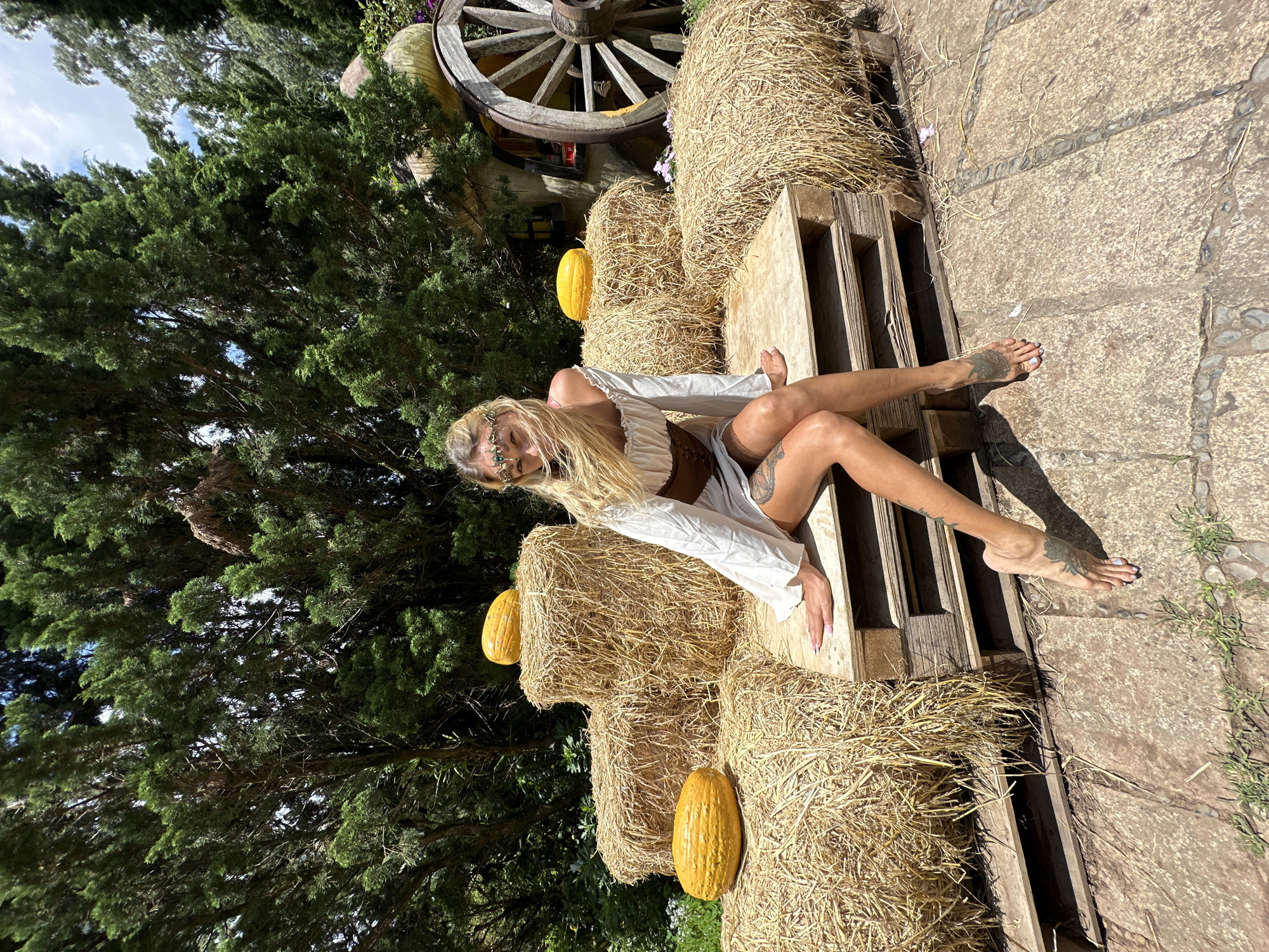 A young woman with blonde hair, wearing a white off-the-shoulder dress and a brown corset, sits on a wooden pallet surrounded by hay bales in an outdoor setting. She has a delicate headdress on her head. She is barefoot with tattoos on her legs. Several yellow gourds are placed on the hay bales. In the background, there are trees and a large wooden wagon wheel. The scene appears to be in a rural or rustic location, possibly part of a harvest festival display. The ground is paved with stone. The lighting suggests it is daytime. This image was taken at the flower garden of Cam Ly in Da Lat, Vietnam.