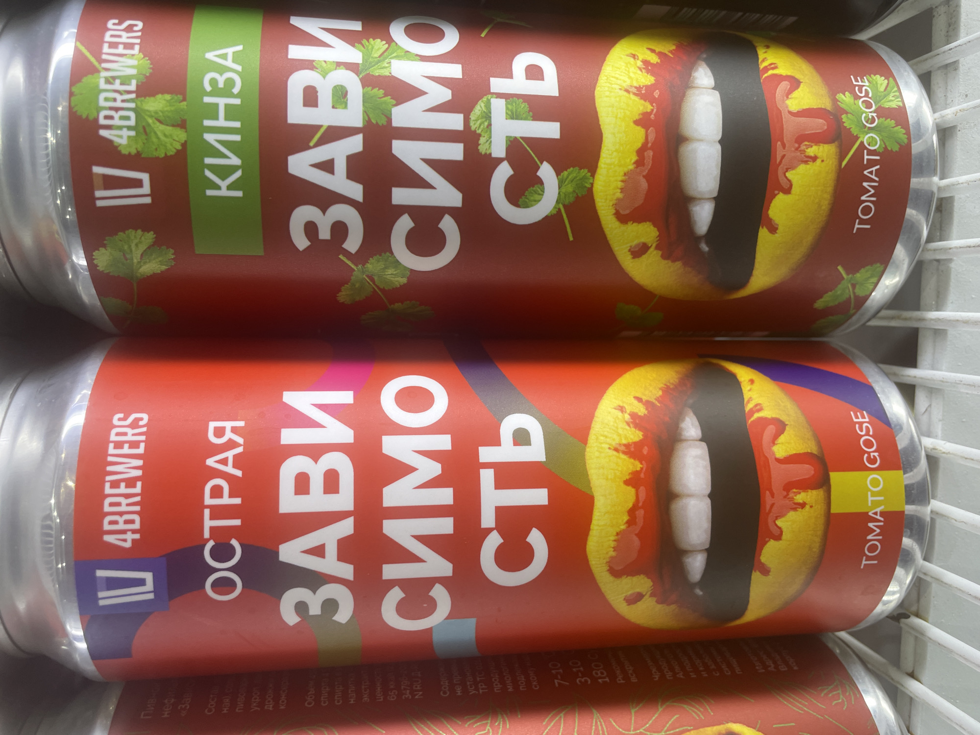 The image shows two cans of beer, likely sitting in a refrigerator or display case. The cans are red with a prominent graphic of a mouth with teeth, splattered with red liquid, resembling blood. Both cans feature large, white Cyrillic text that reads "ЗАВИСИМОСТЬ" (ZAVISIMOST), meaning "addiction." The top can has the word "КИНЗА" (KINZA), meaning "cilantro," on a green background, and the text "TOMATO GOSE." The bottom can has the word "ОСТРАЯ" (OSTRAYA), meaning "spicy," and also reads "TOMATO GOSE." The brand name "IV 4BREWERS" is visible on both cans. The scene appears to be indoors, with the cold, metallic structure of the display case visible. The lighting suggests it could be daytime, possibly in a store or bar. The overall aesthetic is bold and somewhat provocative, with the "addiction" theme and graphic imagery.