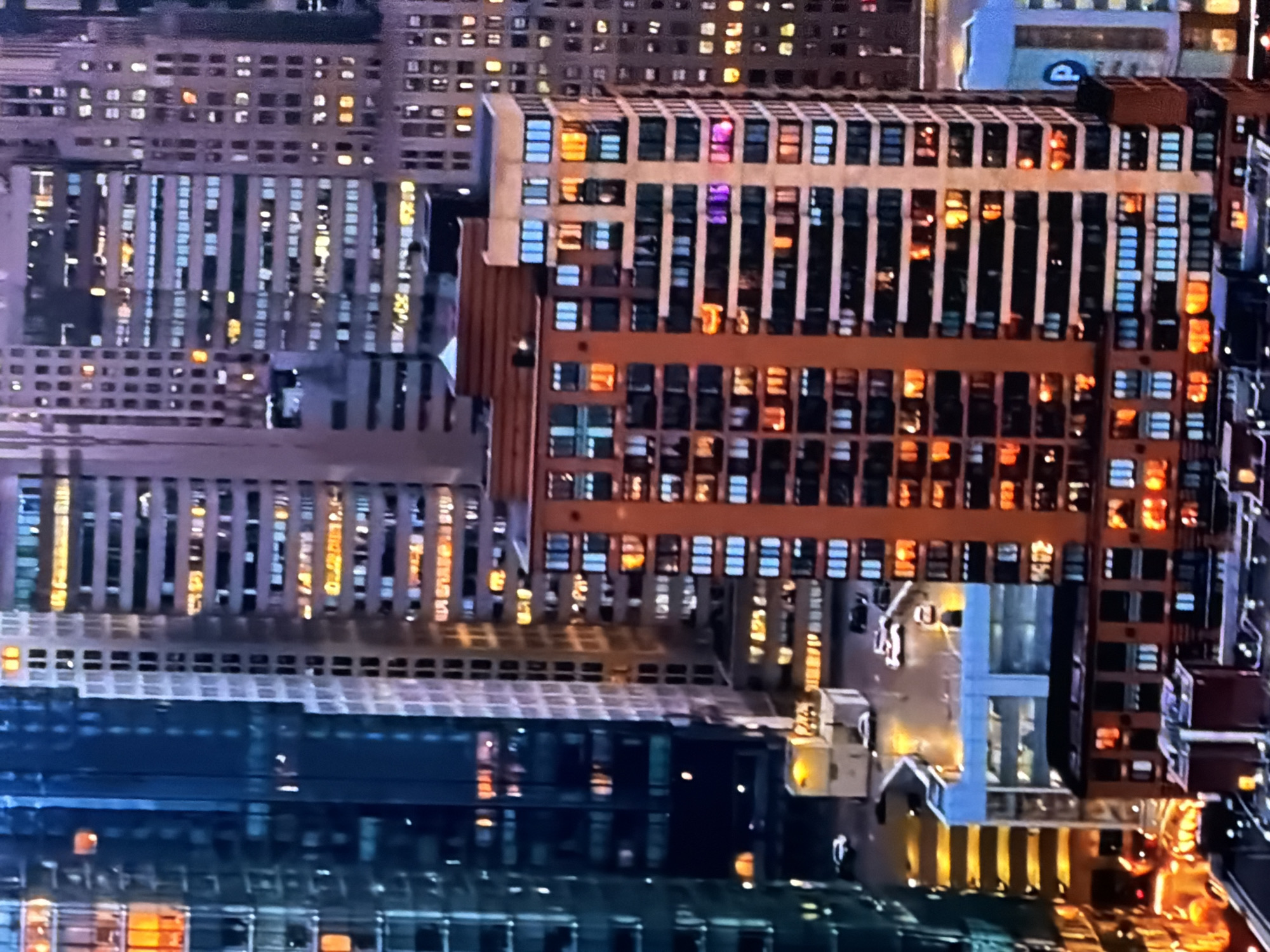 The image displays a dense urban cityscape at night or dusk, located in the city of Maidstone, United Kingdom. The scene is observed from an elevated viewpoint, likely from a tall building, looking across a concentration of high-rise structures. On the left, a dark, modern building with a blue-green glass facade reflects ambient light, with several internal windows illuminated in warm orange and yellow tones. To its right and in the background, numerous multi-story buildings of varying heights and architectural styles feature hundreds of windows, a significant portion of which are lit, casting warm yellow, white, and occasional blue lights. In the foreground-center, a prominent reddish-brown building, appearing to be residential or mixed-use, displays a grid of uniformly spaced windows. Many of these windows are illuminated with warm orange light, while some on the upper levels show distinct blue and magenta or purple light. Below the primary buildings, sections of ground level are visible, showing what appear to be streets or parking lots with several vehicles, primarily white and dark sedans, either parked or in motion. Small ground-level structures and illuminated streetlights are also present. A blue sign with a white 'P' symbol, indicative of a parking facility, is visible on the bottom-right. No discernible people, direct actions, or interactions are visible within the frame.