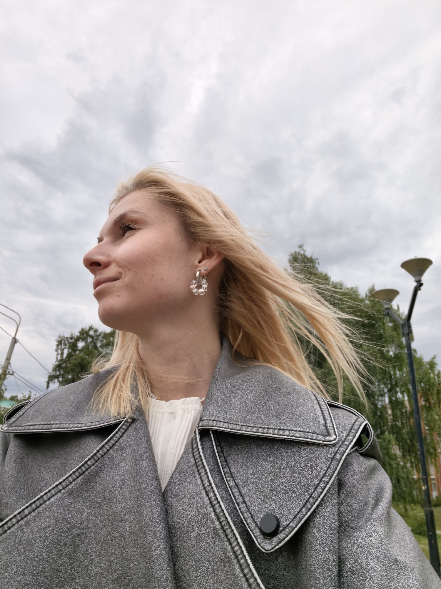 A young woman with blonde hair is pictured from the chest up, looking to her left. She is wearing a grey leather jacket and a white top. She has a large hoop earring with clear beads in her left ear. Her hair is blowing in the wind. In the background, there are trees, streetlights, and a cloudy sky. The location is implied to be Sterlitamak, Russia.