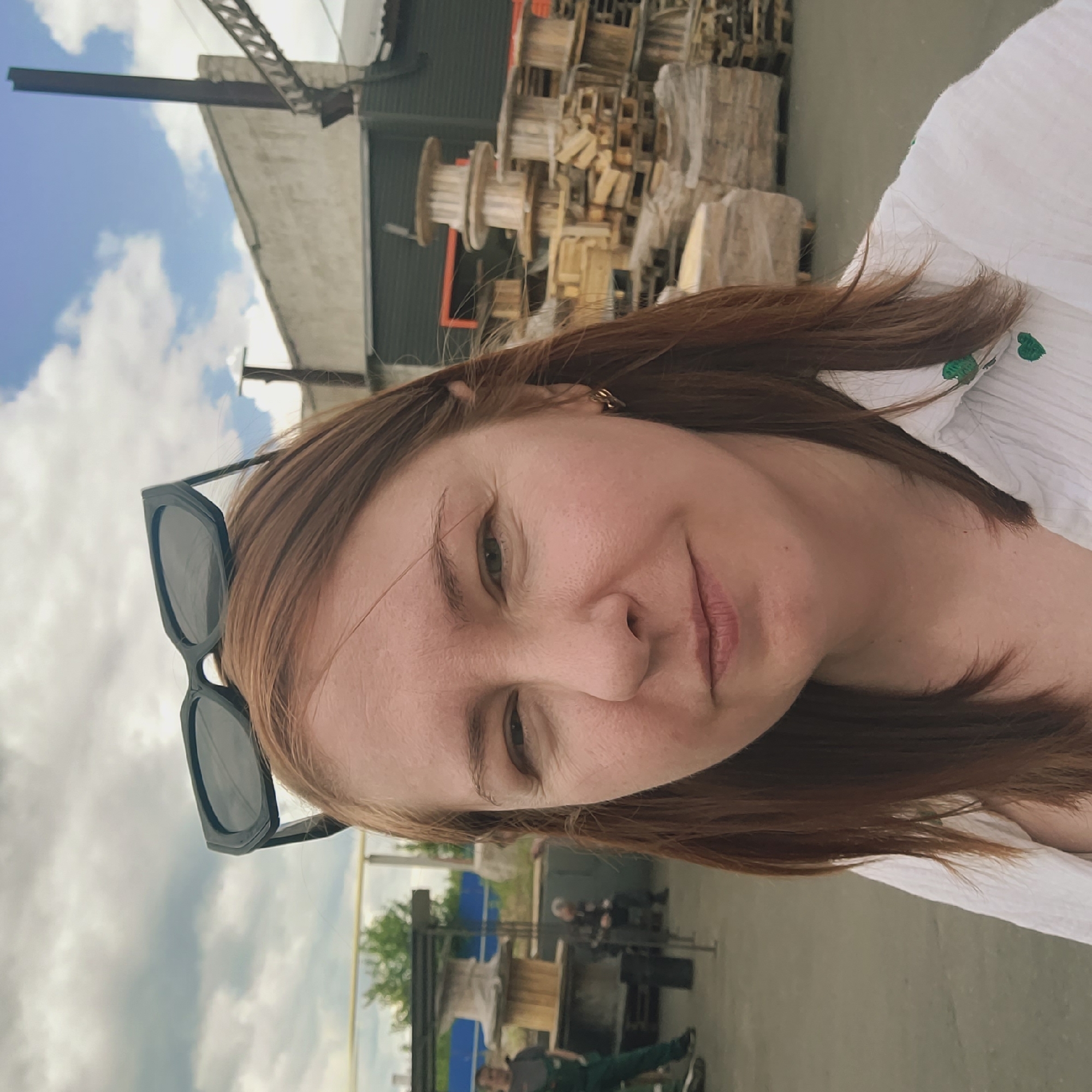 A woman is featured in a close-up, slightly tilted selfie, looking directly at the camera with a relaxed expression and a subtle smile. She has reddish-brown hair, with black sunglasses pushed up onto her head. She is wearing a white shirt with small green embroidered details. A golden earring is visible on her right ear.
The background suggests an outdoor, industrial or storage yard setting during the daytime under a cloudy sky. Prominently visible are piles of wooden pallets and large wooden spools or reels. Behind these, there are dark-colored buildings or structures. In the far distance, blurred figures of other people and blue barrel-like objects can be discerned. The overall impression is a casual portrait taken in an industrial or work-related environment. No text is visible in the image.