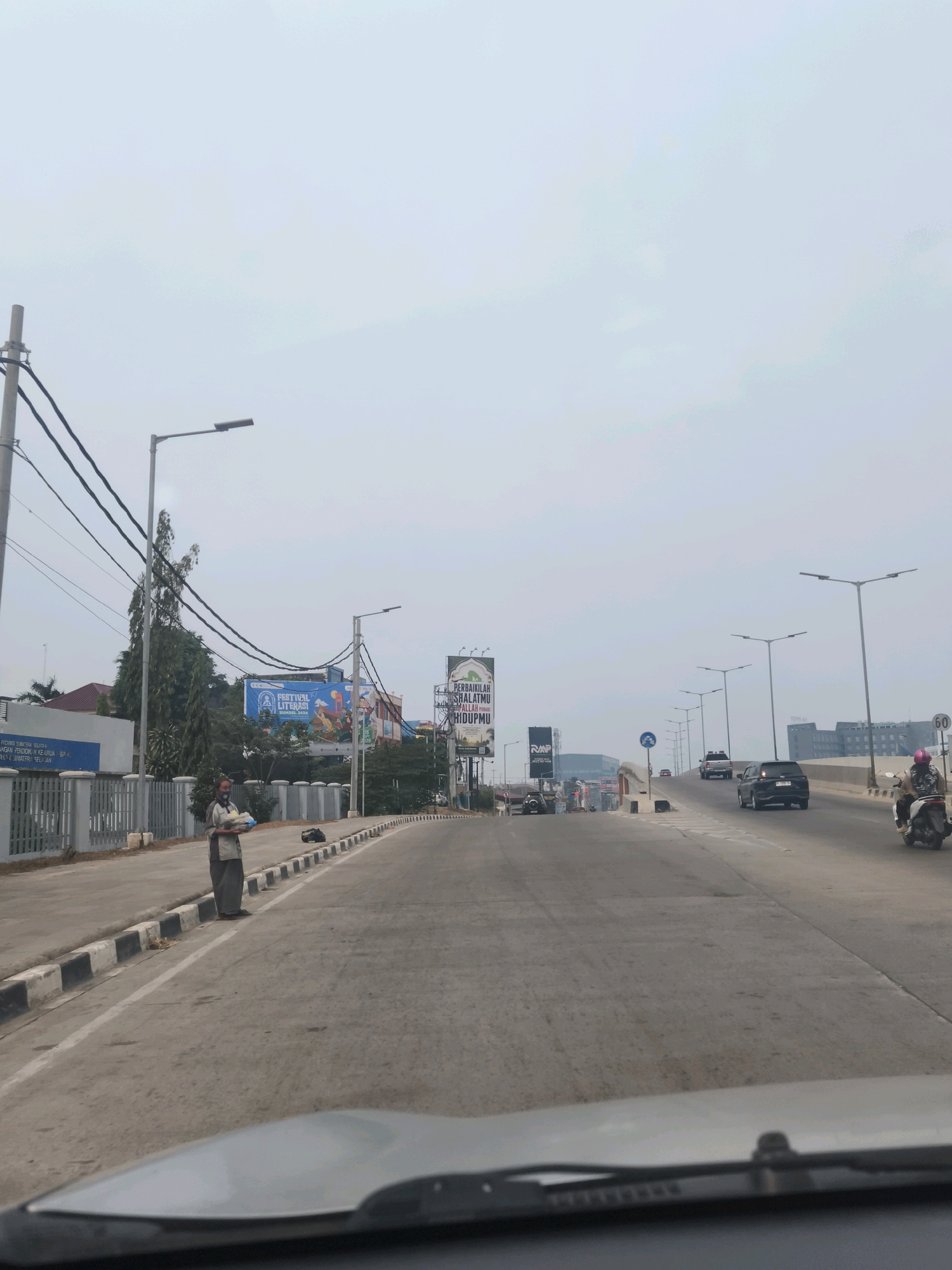 A medium-shot image captured from a moving vehicle shows a street scene in Palembang, Indonesia, on a bright, overcast day. The road is paved, with a distinct curb and sidewalk on the left. A person wearing a mask and traditional clothing stands on the sidewalk, holding what appears to be a package of food or goods. Their presence is notable against the backdrop of urban infrastructure.
The environment includes several streetlights, power lines crisscrossing the sky, and a few trees along the sidewalk. Several vehicles, including cars and a motorcycle with a rider, are visible on the road, indicating moderate traffic. Billboards are also present, with one displaying text in what appears to be Arabic script and another with graphics related to a "Festival Literasi."
The architecture in the background includes buildings of various sizes, some with modern facades and others appearing more industrial. The sky is a pale blue with diffused clouds. The perspective suggests the image was taken from the driver's or passenger's seat of a car, looking forward along the road. The overall impression is of a typical urban street on a day with less than ideal weather conditions.