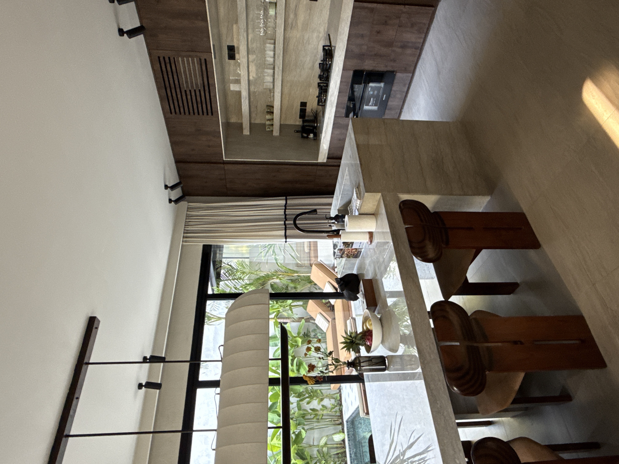 The image shows an interior space in Kuta Selatan, Indonesia. A kitchen is visible, featuring dark brown cabinetry, a light-colored countertop, a stovetop, and storage for glassware. The space transitions to a dining area with a light-colored countertop island and wooden chairs. A window reveals an outdoor area with greenery. A decorative light fixture stands near the window. No people are visible.