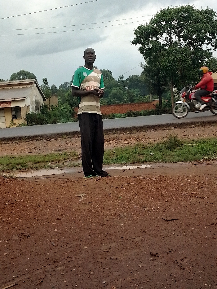 A lone man in a green and white striped t-shirt and black pants stands in a dirt lot in Kivaywa, Kenya. He stands facing the camera with his hands clasped in front of him. The man is standing to the right of a paved road. A motorcycle is visible in the background, heading in the direction of the man. There are two buildings visible in the background, as well as several trees. The sky is overcast and a small puddle of water is visible in the dirt lot. The coordinates (latitude: 0.56, longitude: 34.806) place the location in the town of Kivaywa, Kenya.