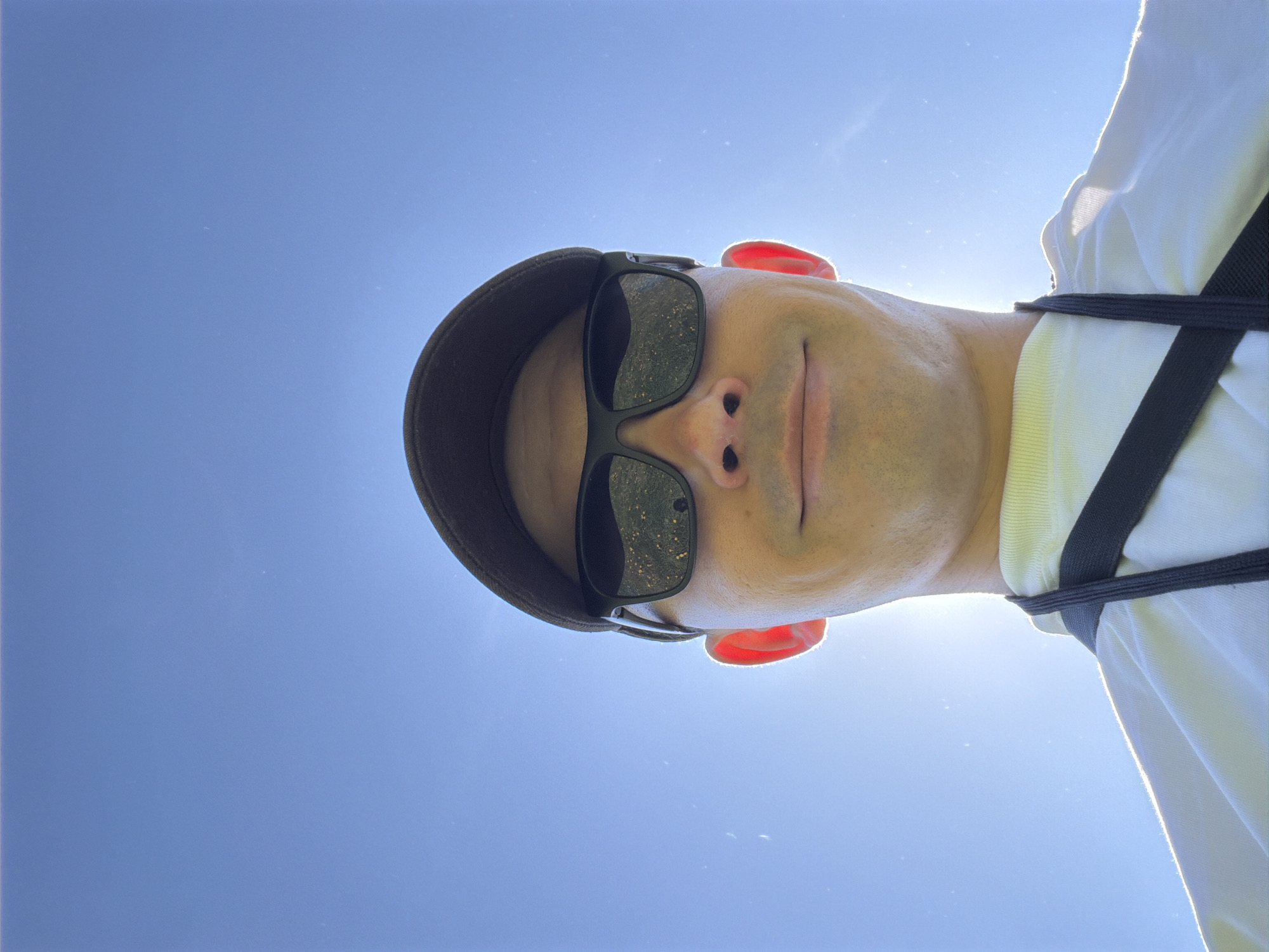 A low-angle close-up shot of a person wearing a baseball cap and sunglasses, set against a clear blue sky. The person appears to be wearing a white shirt with a black harness strap across the chest. The reflection in the sunglasses lenses is indistinct, but it suggests an outdoor environment. The skin tone is light, with noticeable redness in the ears. The photograph does not provide enough information to confirm the location is Oreshniki, Russia. No specific actions or interactions are visible.