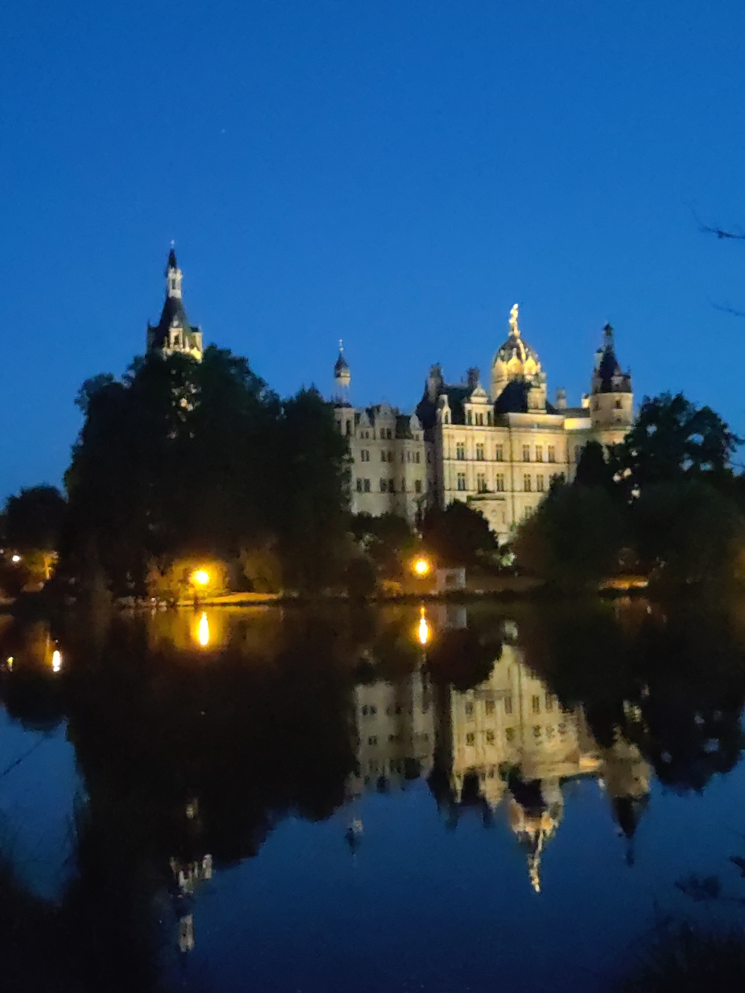 This is a nighttime, outdoor, full-frame photograph of the Schwerin Castle, located on an island in the city of Schwerin, Germany. The castle, illuminated by exterior lights, is a large, multi-turreted building with a golden dome. It is surrounded by trees and reflected in the dark water of a lake in the foreground. The sky is a deep blue, indicative of twilight or early night. The reflection of the castle and the surrounding lights creates a mirrored image in the water. There are no people or animals visible in the scene.