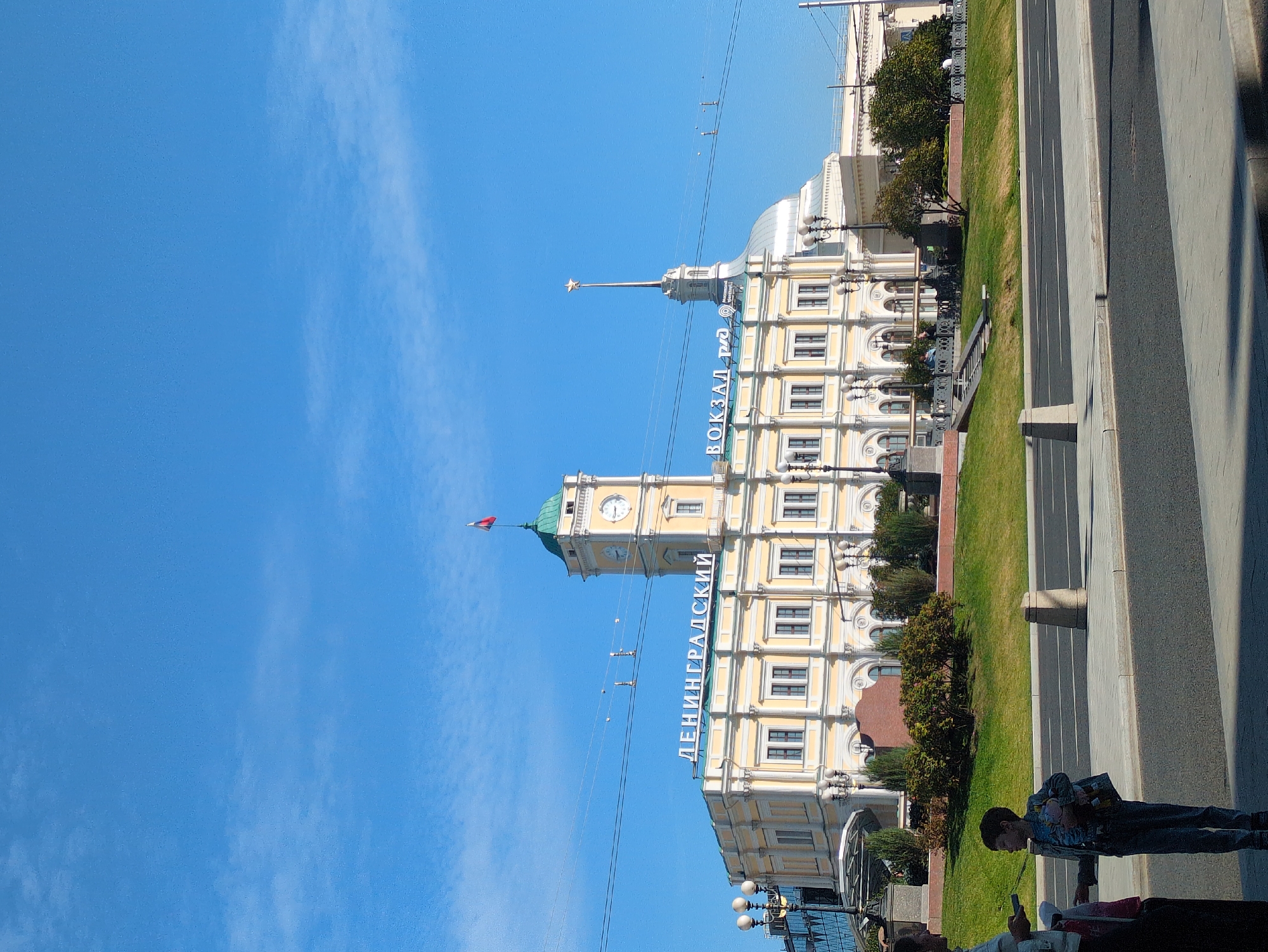 The image shows the Leningradsky railway station in Moscow, Russia, on a clear day. The station building is a large, yellow, neoclassical structure with a prominent clock tower. The words "ЛЕНИНГРАДСКИЙ" (Leningradsky) and "ВОКЗАЛ.РФ" (Vokzal.RF) are visible on the facade. A Russian flag flies from the top of the clock tower. In the foreground, a young boy wearing a denim jacket and jeans stands holding a toy pig and a bag. He is facing towards the left of the frame, looking at a woman who is partially visible on the left edge of the image. They appear to be standing on a sidewalk next to a road. The area around the station is landscaped with green grass and trees.