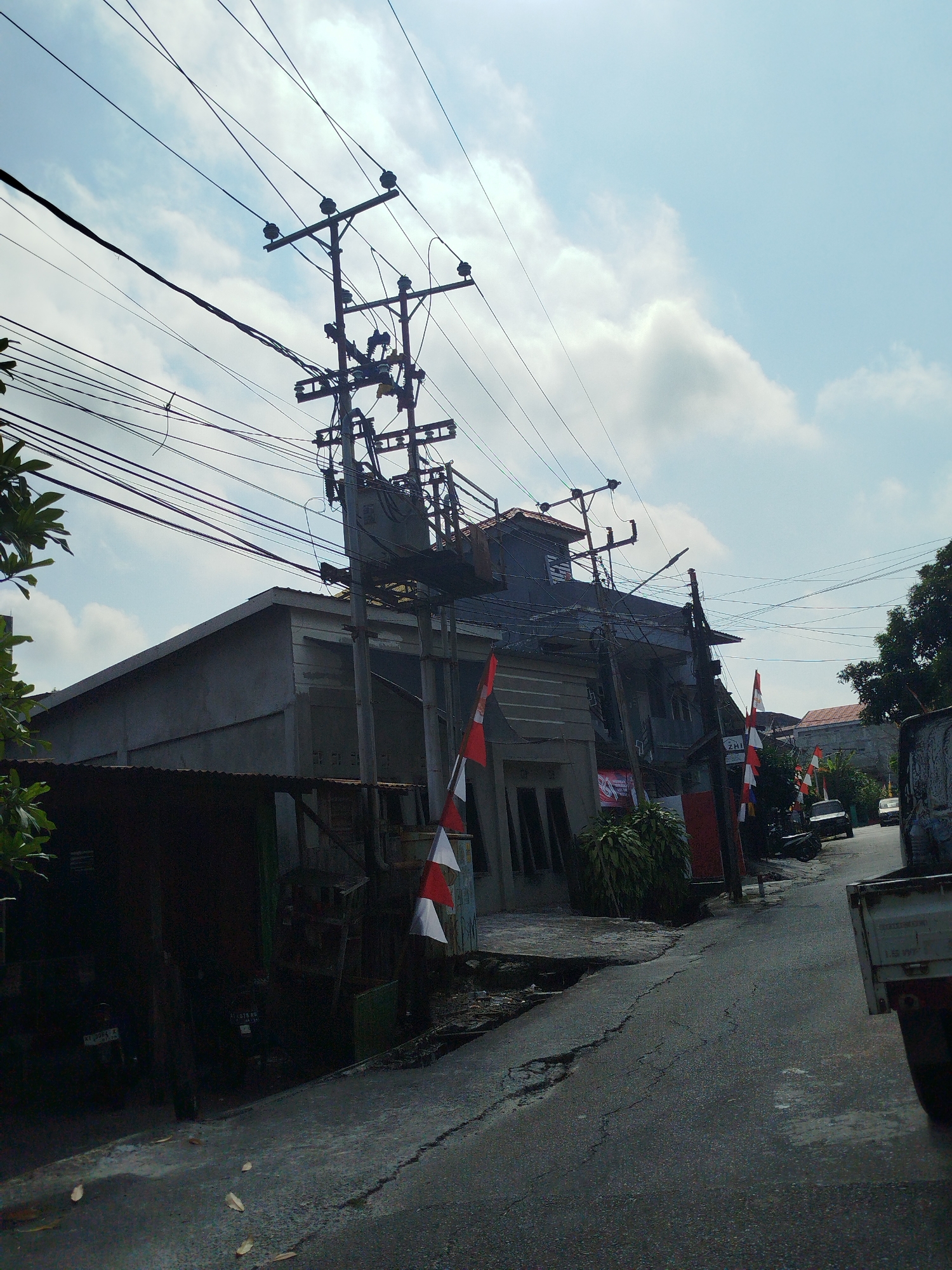 The image shows a street scene in Balikpapan, Indonesia, dominated by an overhead electrical utility pole and complex network of wires. The pole is situated on the right side of the street, with a transformer visible. Several buildings line the street, appearing to be a mix of residential and commercial structures, some with corrugated metal roofs. Small Indonesian flags, red and white, are displayed in front of some of the buildings. The street itself is asphalt and shows signs of wear and tear, with cracks and uneven surfaces. There are a few parked vehicles visible, including a white pickup truck on the right side of the frame and a dark-colored car further down the street. The sky is partly cloudy with a bright, diffused light, suggesting daytime. Trees and foliage are visible on the left and right edges of the frame. The overall impression is of a typical urban street in Indonesia, with infrastructure and daily life elements captured in the photograph.