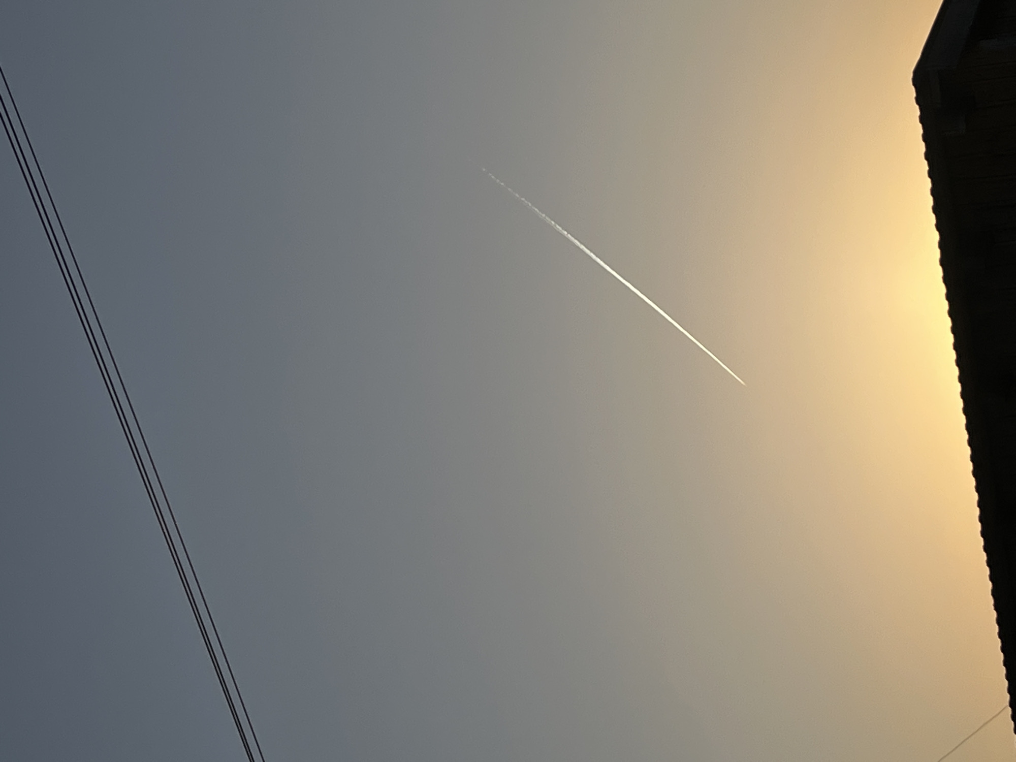 The image captures an outdoor sky scene in the city of Фоки, Russia, during either sunrise or sunset. The sky transitions from a muted blue-gray in the upper portion to a prominent golden-orange hue near the horizon at the bottom. A single, linear, white contrail from an aircraft extends diagonally from the lower-left quadrant towards the upper-right. Two parallel, dark-colored utility wires are visible in the upper-left portion of the frame, running diagonally. The silhouette of a building's roofline, featuring what appears to be a textured or tiled surface and a distinct corner, occupies the bottom edge of the image, primarily in the lower-right. No individuals, specific actions, or interactions are visible.