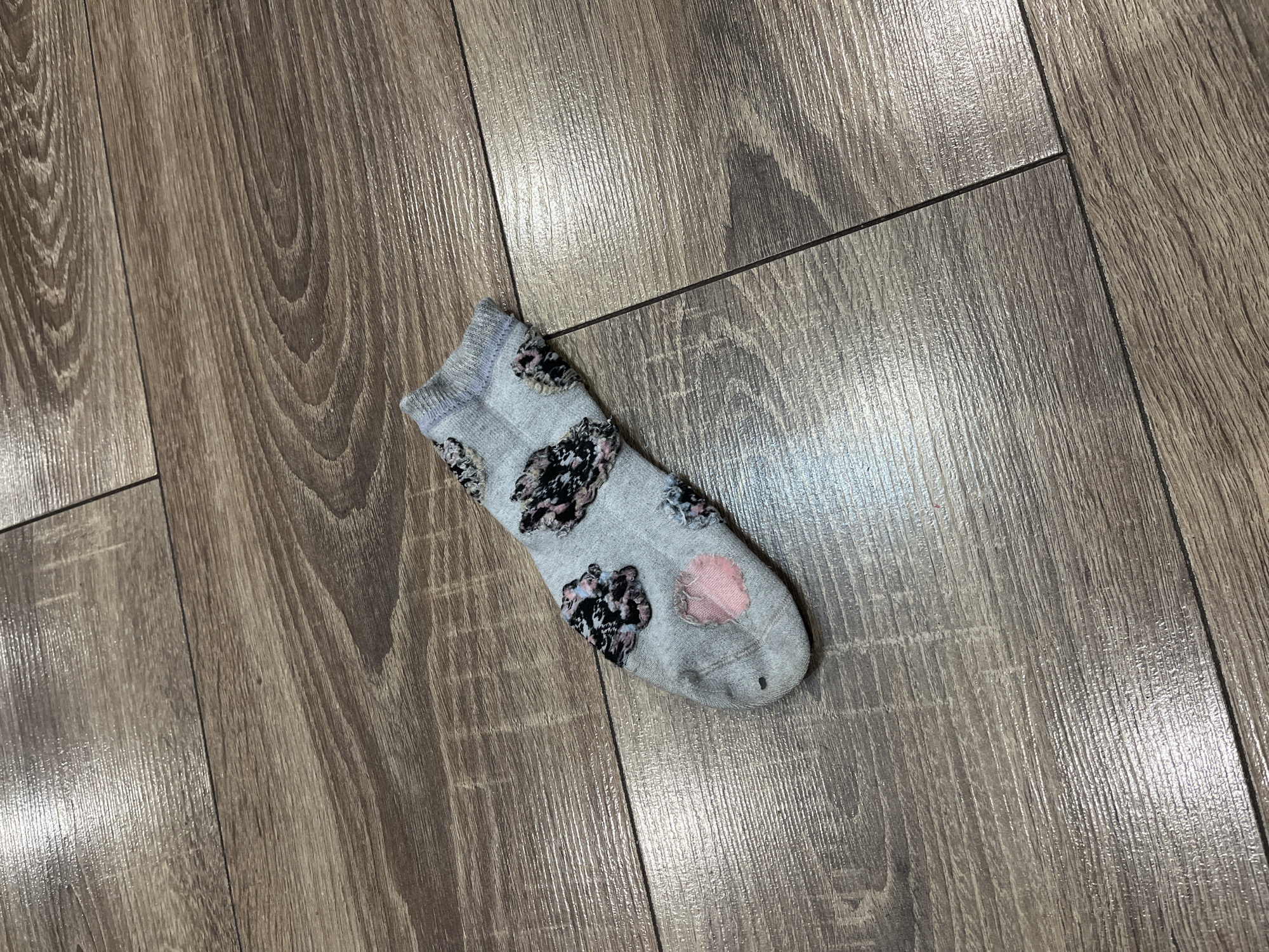 A single gray sock with a floral pattern is lying on a wooden floor. The sock is angled from the bottom left to the top right of the frame. The floor appears to be laminate flooring with a wood grain texture. There are no people or other objects visible in the image. The scene is indoors, likely in a residential setting. There is no information to suggest the location is Rossiiskii, Russia.